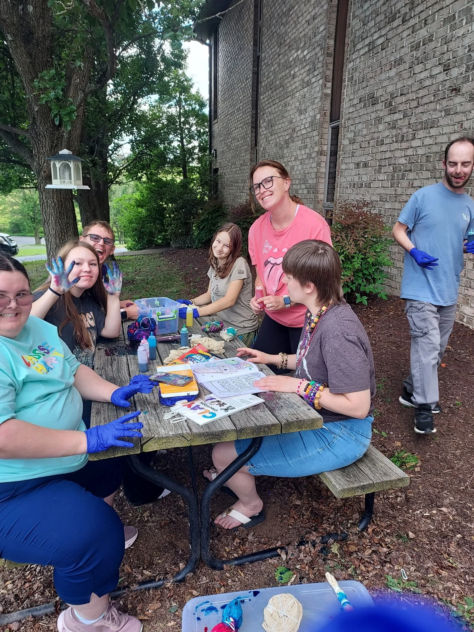 A group of people are gathered around an outdoor wooden picnic table participating in a tie-dye craft activity next to a brick building. Several individuals sit at the table wearing blue protective gloves, with tie-dye bottles, rubber-banded shirts, 