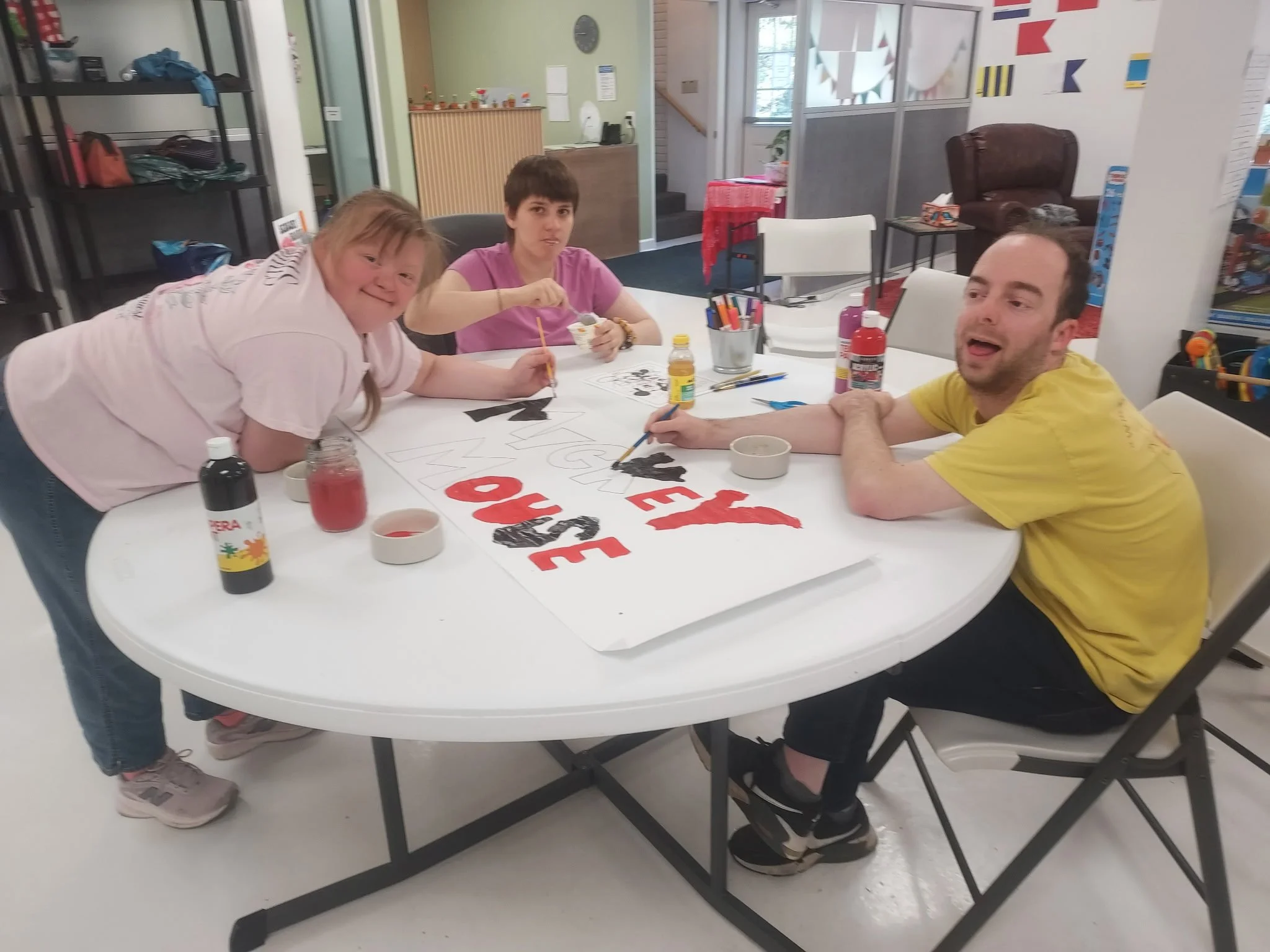 Three people are gathered around a large, white circular table inside a brightly lit activity room, painting a large sign. On the left, a woman in a light pink t-shirt and jeans leans over the table, smiling toward the camera. In the center, a woman 