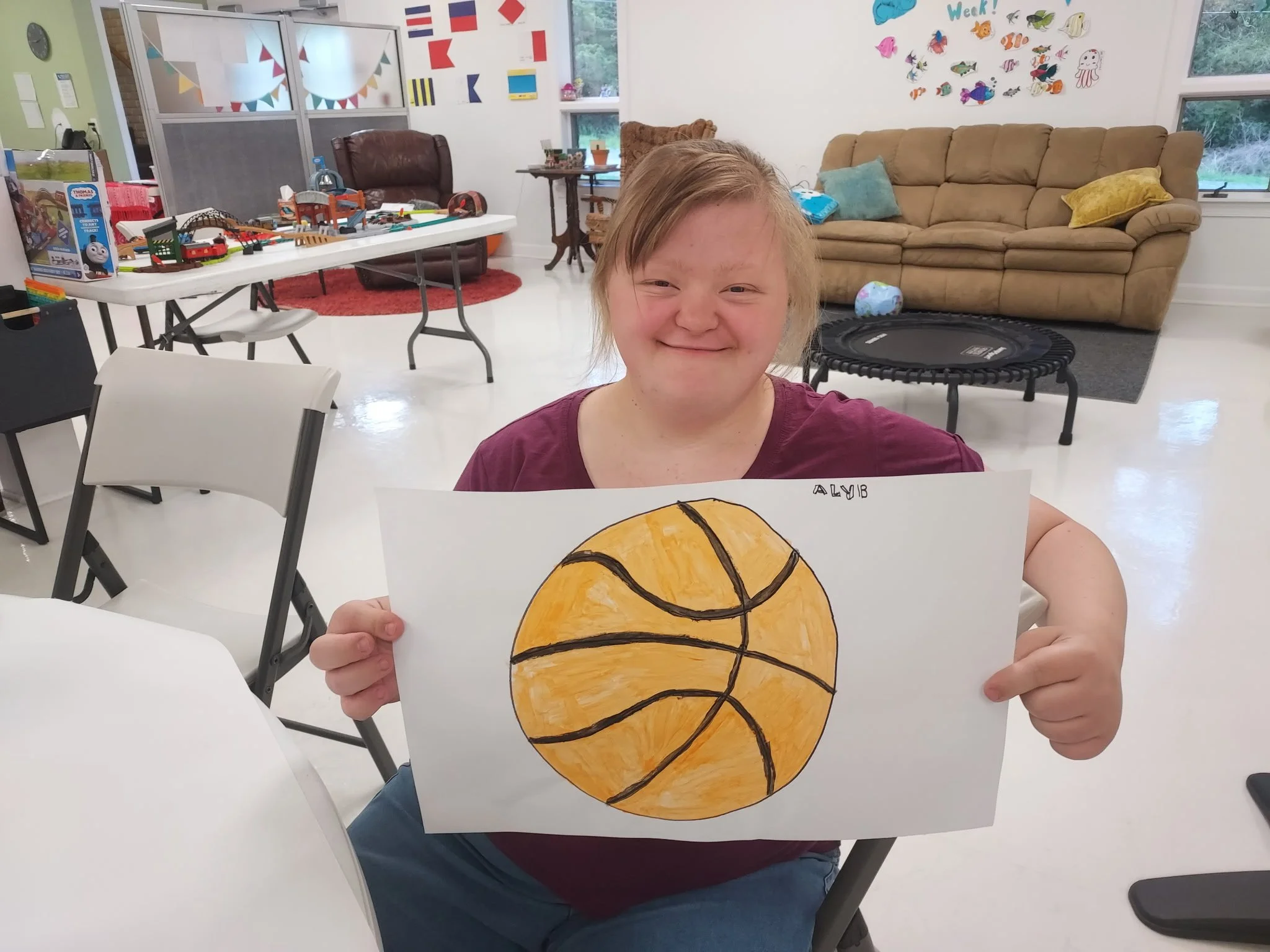 A woman sits in a white folding chair inside a bright community room, smiling as she holds up a white piece of paper. On the paper is a large, hand-drawn and painted orange basketball with black lines, signed "ALY B" in the upper right corner. She is