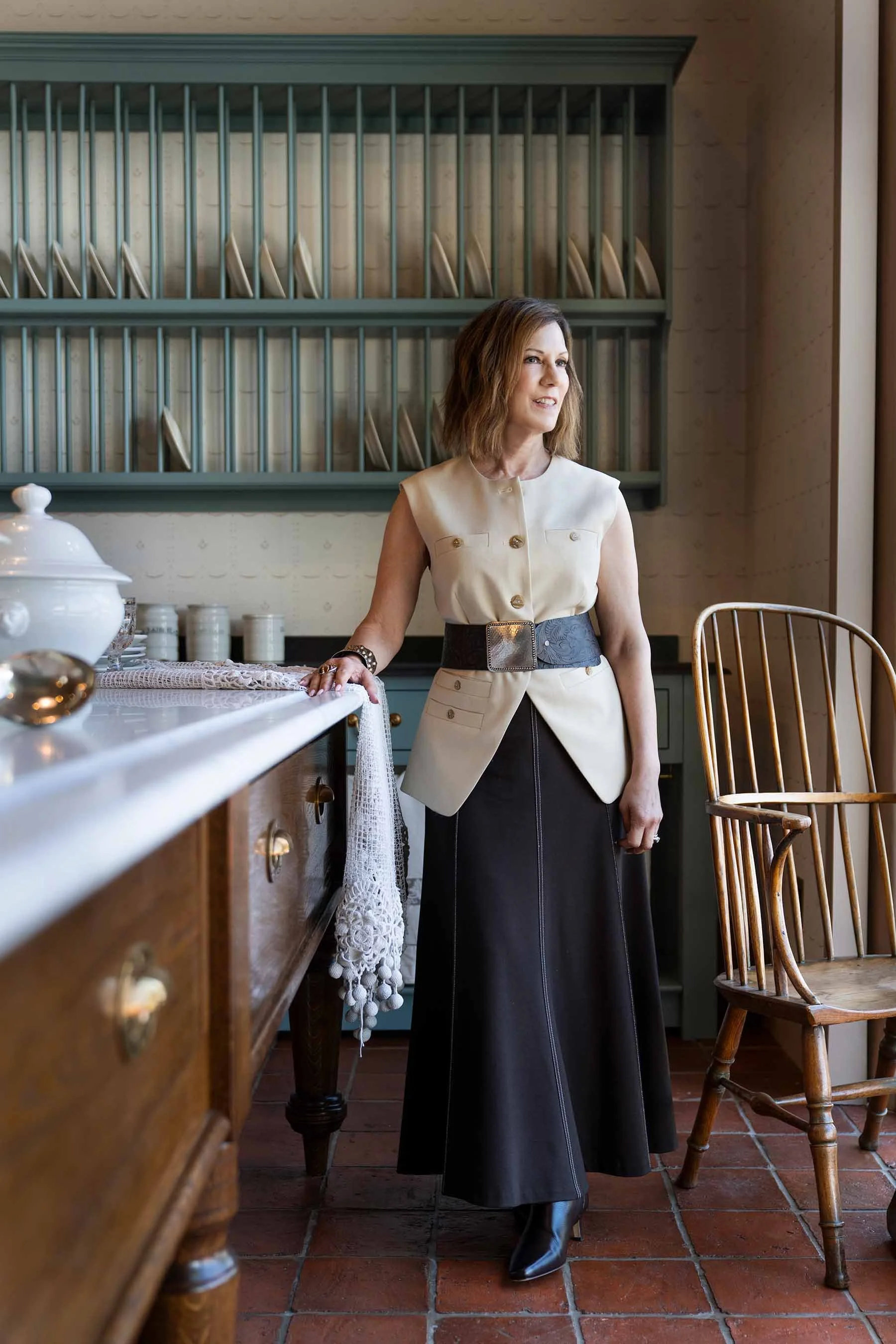 Janet T. Marena standing in a sage-green kitchen with a vintage plate rack and terracotta tile floor