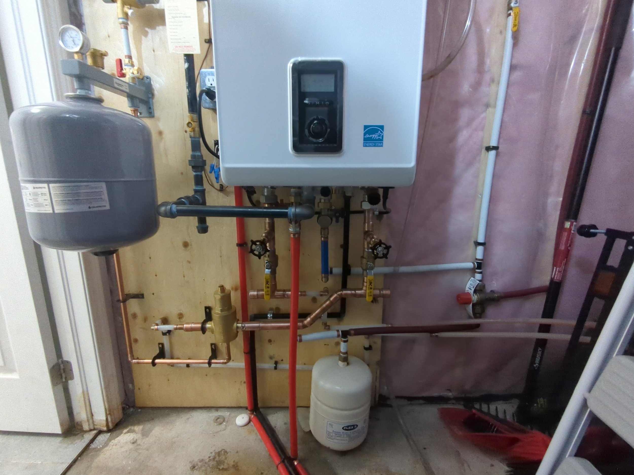 A boiler system with various pipes, valves, and a pressure tank installed on a small wall in a utility room.