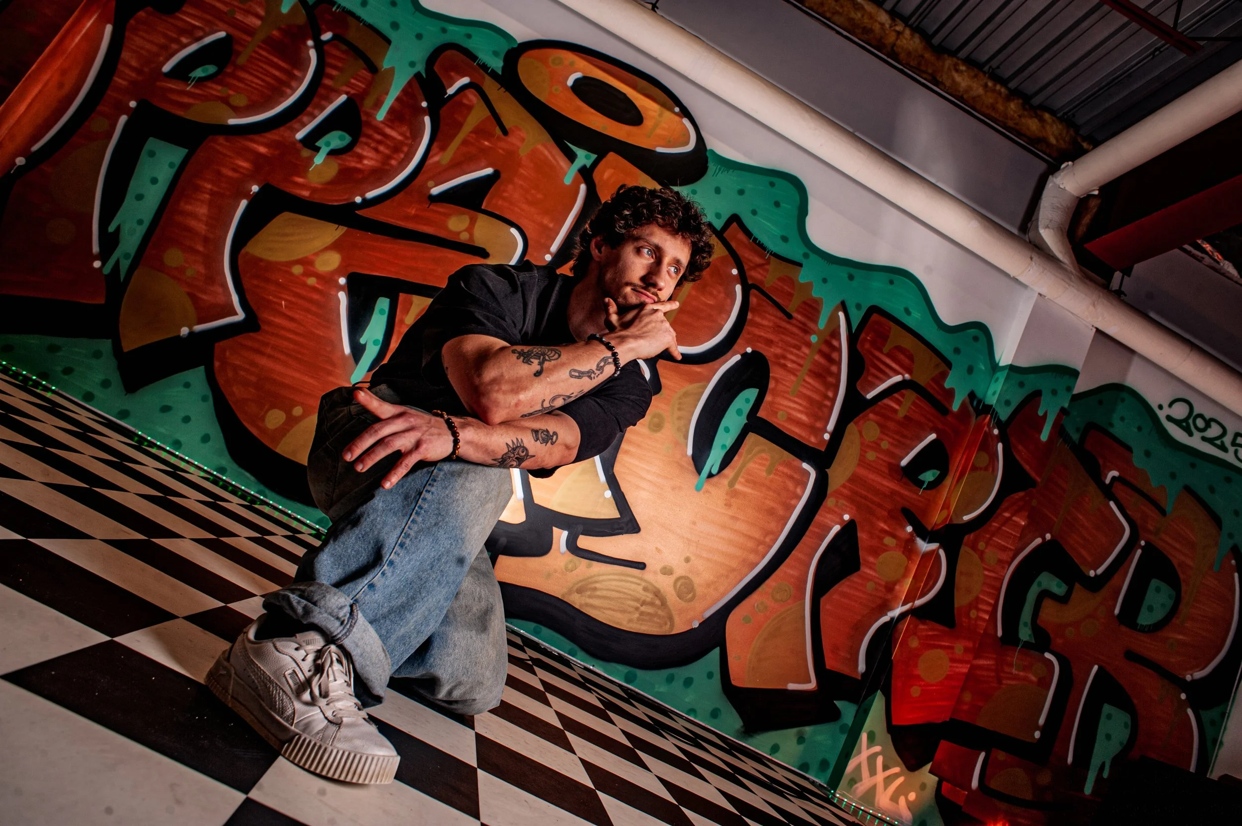 Un homme avec des tatouages, vêtu d'un t-shirt noir, jeans et baskets blanches, pose en kneeling devant un mur de graffiti coloré dans un endroit avec un sol en carreaux noir et blanc.