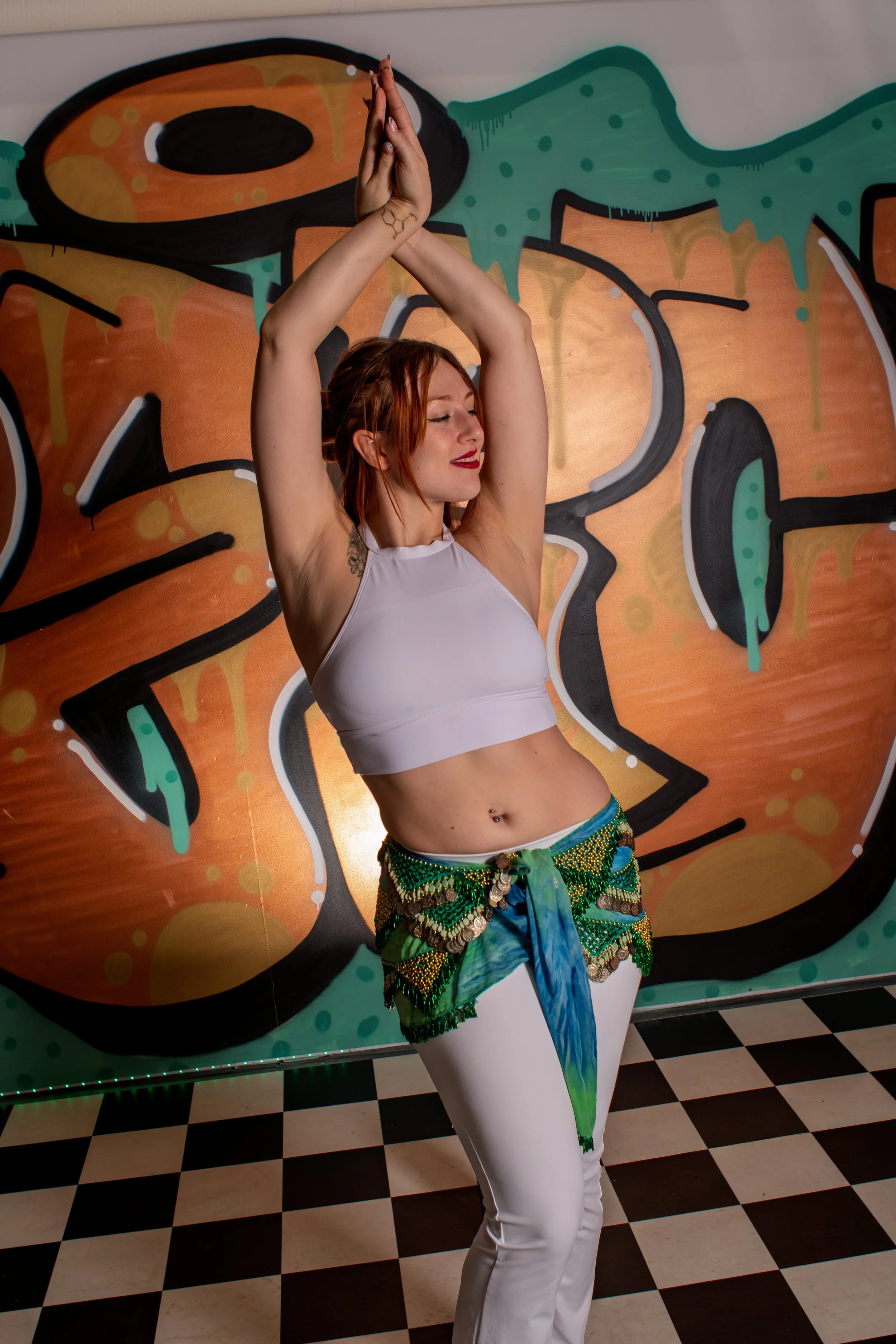 Une femme danse avec les bras levés au-dessus de sa tête devant un mur graffité coloré, portant un haut blanc, un pantalon blanc et une ceinture décorative verte avec des pièces métalliques, sur un sol à carreaux noir et blanc.