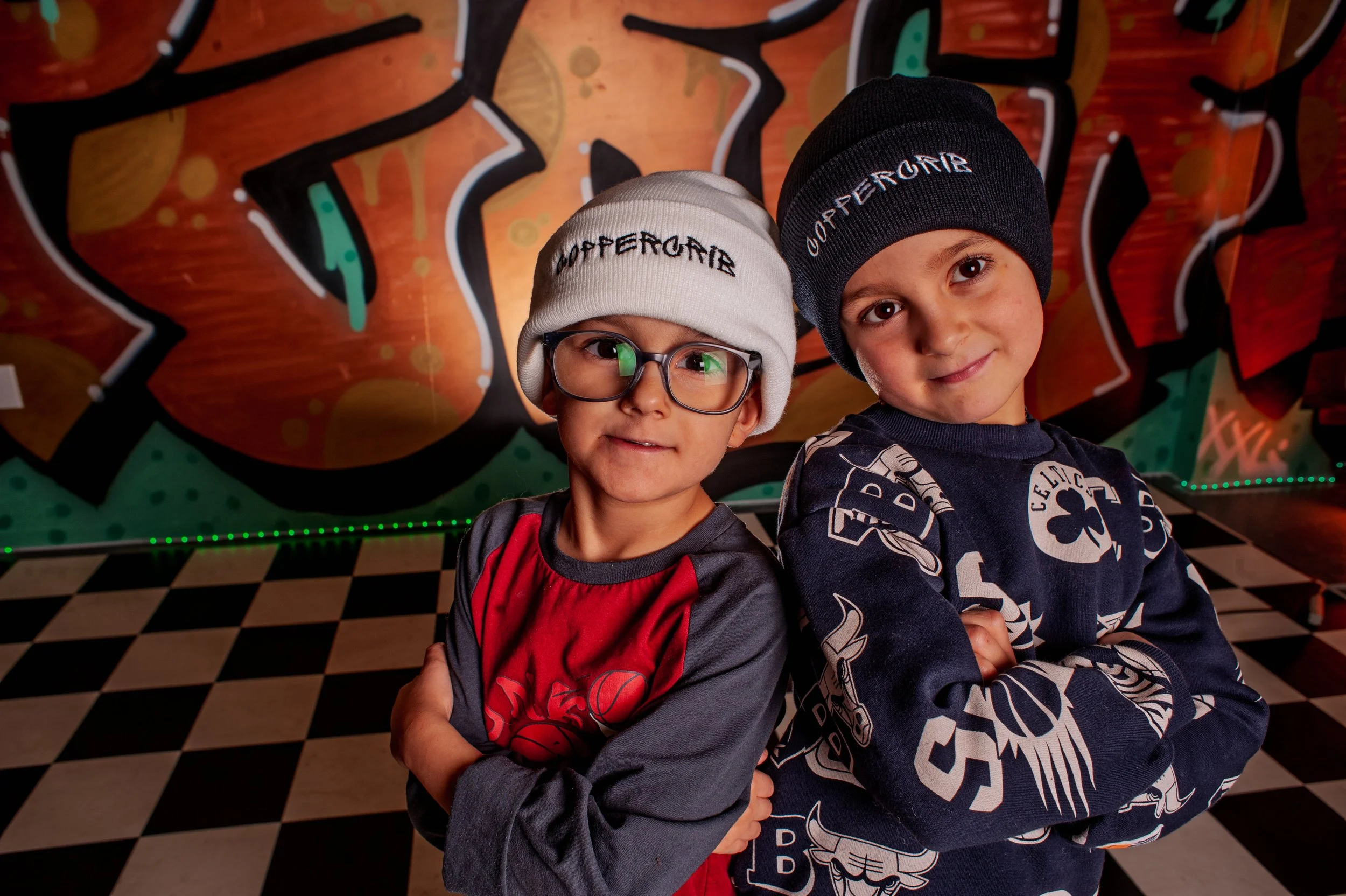 Deux jeunes enfants posent de manière confiante avec des bras croisés devant un mur graffiti coloré. L'un porte des lunettes, un bonnet noir, et un sweat à motifs de cartes de jeu. L'autre porte un bonnet blanc et un sweat à manches longues gris et rouge.