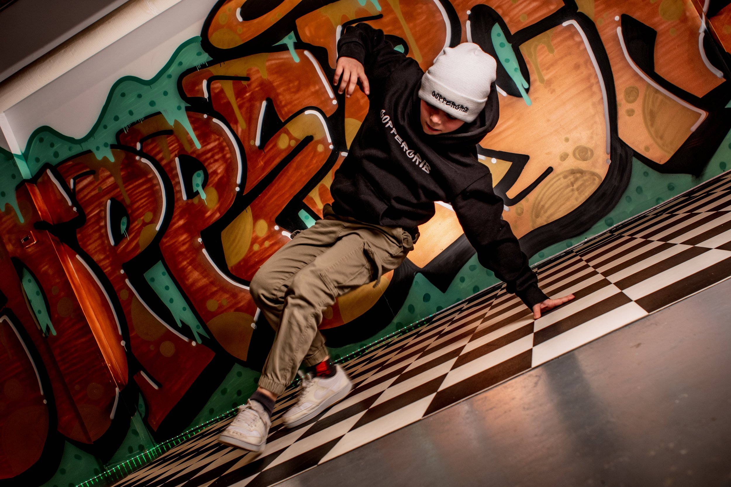 Un garçon en hoodie noir, pantalon beige, et bonnet blanc fait du breakdance devant un mur coloré de graffiti avec un sol en motif damier.