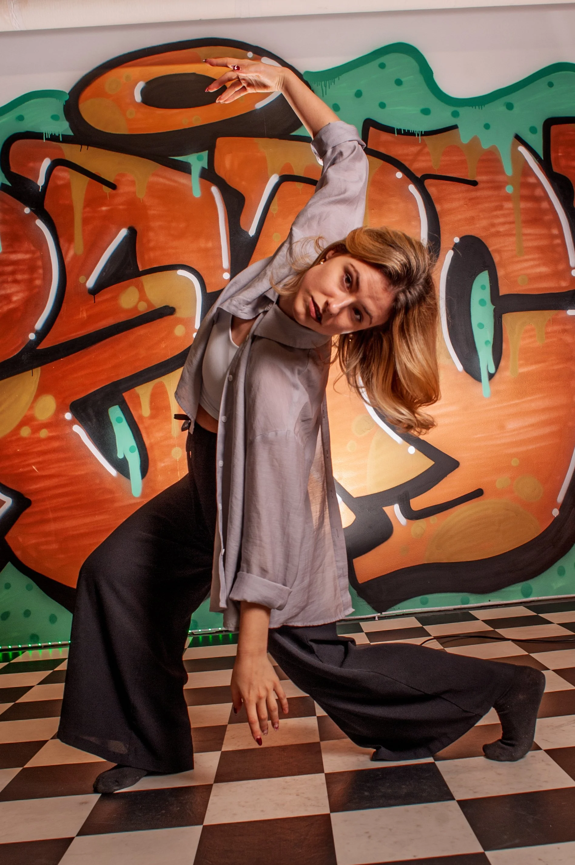 Une jeune femme pose devant un mur avec graffiti coloré. Elle porte un chemisier gris, un débardeur blanc et un pantalon ample. Elle a les cheveux blonds et roux et adopte une posture artistique en inclinant la tête et en levant un bras.