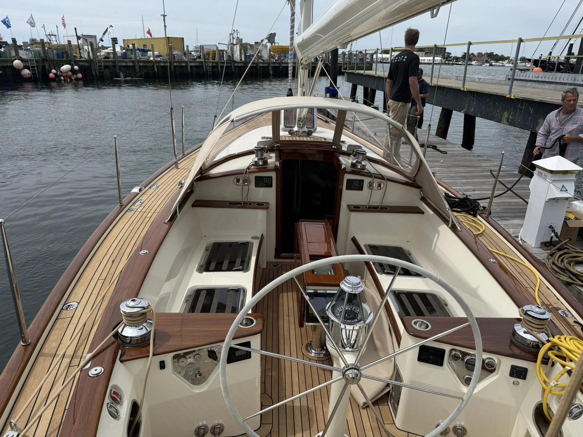 View of a classic sailboat docked at a marina pier with people nearby, showing the boat's interior and steering wheel.