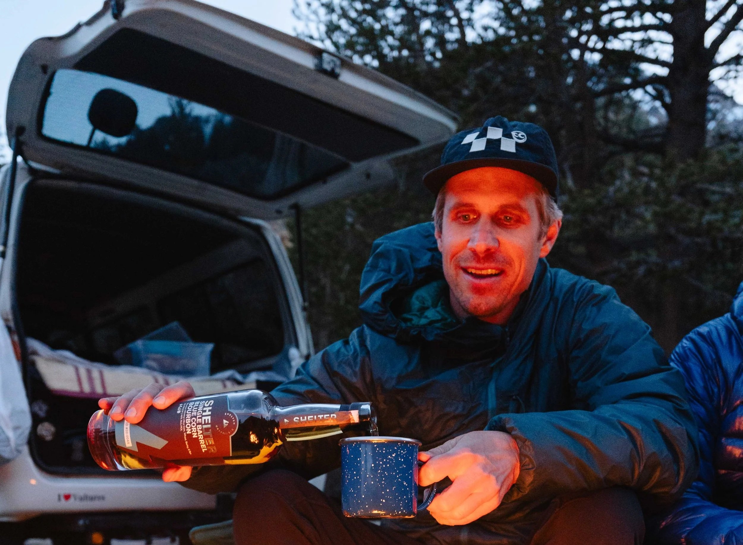 A man in outdoor gear is pouring whiskey from a bottle into a mug while sitting outside near an open vehicle trunk at dusk or early evening.