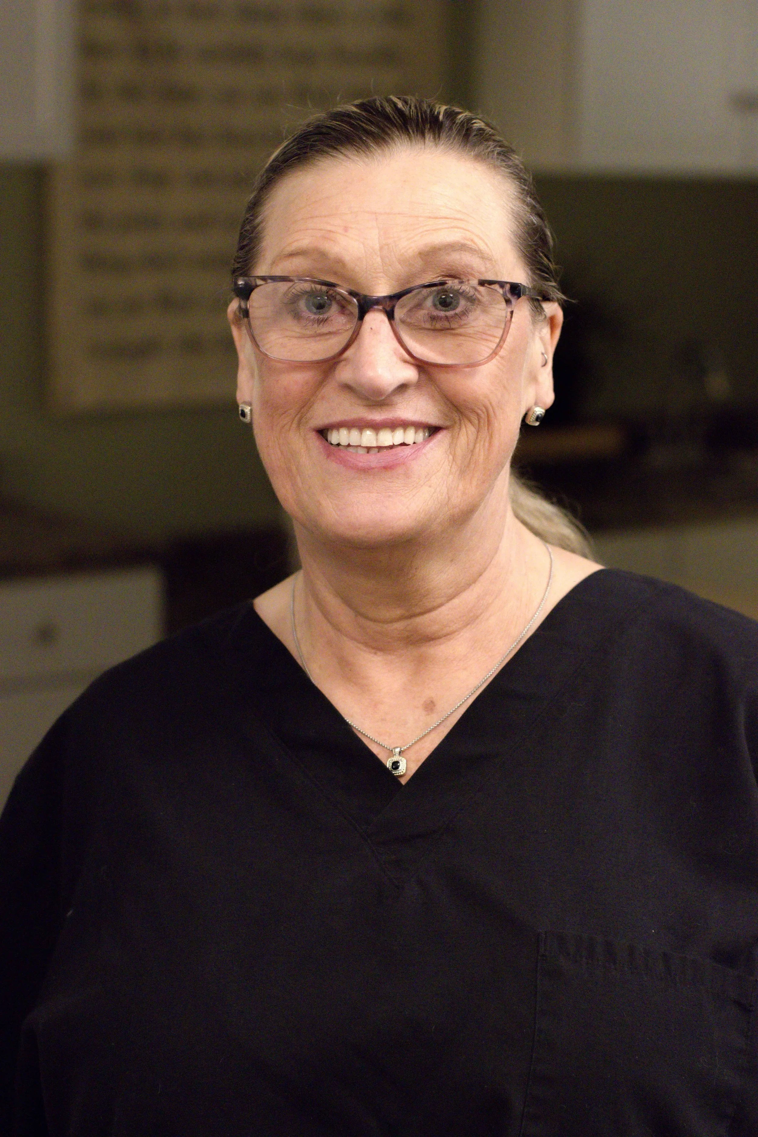 A middle-aged woman with glasses, earrings, and necklace smiling in a black shirt.