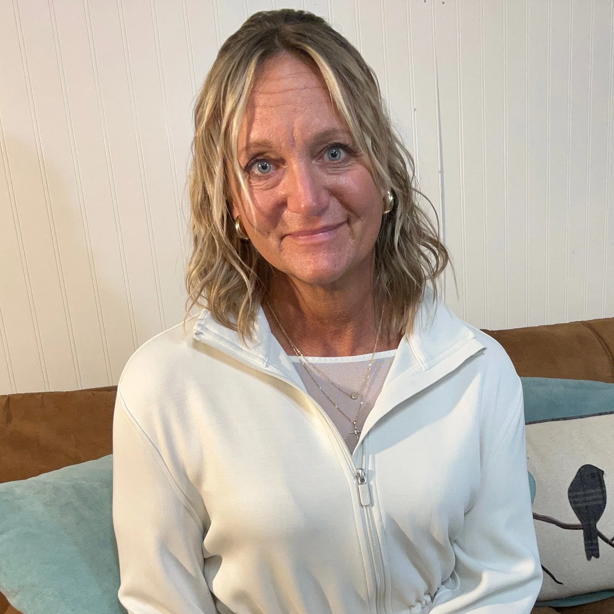 A smiling woman with shoulder-length blonde hair, wearing a white zip-up jacket and layered necklaces, sitting on a sofa in a room with white paneled walls.
