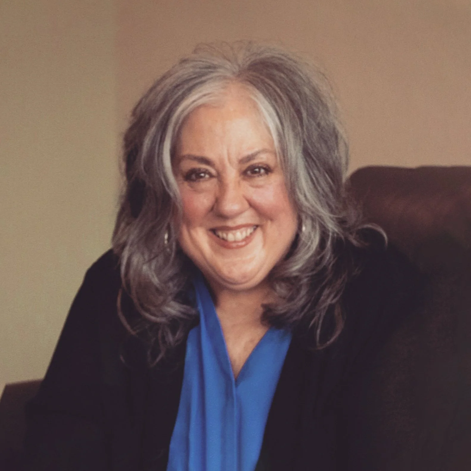 A smiling middle-aged woman with gray and dark hair, wearing a black blazer and blue blouse, sitting on a dark-colored couch.
