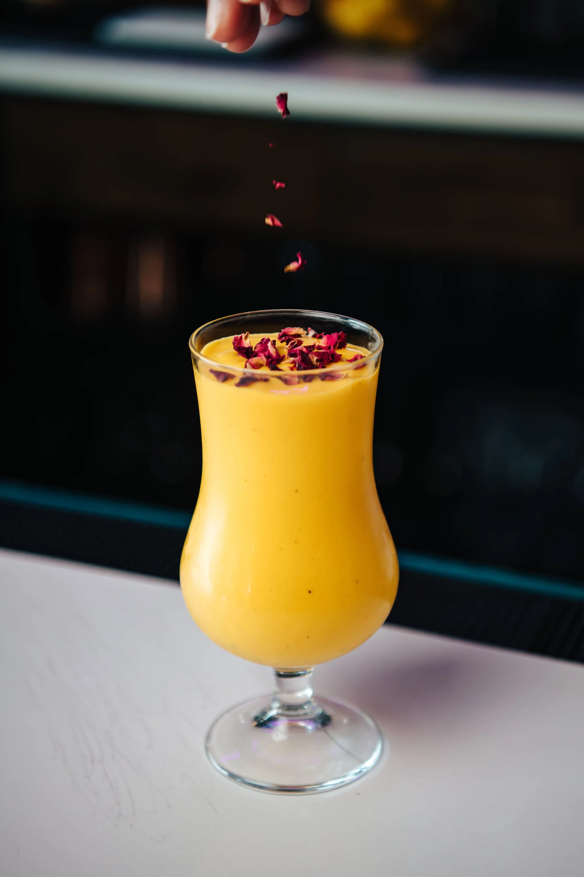 A yellow smoothie topped with dried rose petals in a tall, curved glass on a white surface.