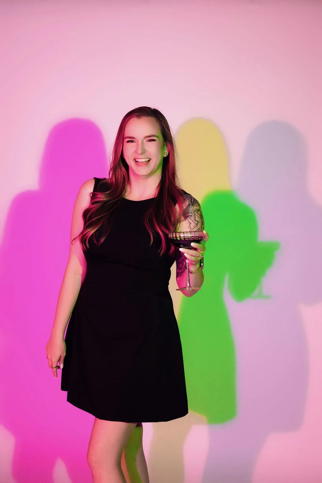 A young woman with long pink hair, smiling and holding an espresso martini, in front of a technicolor background.