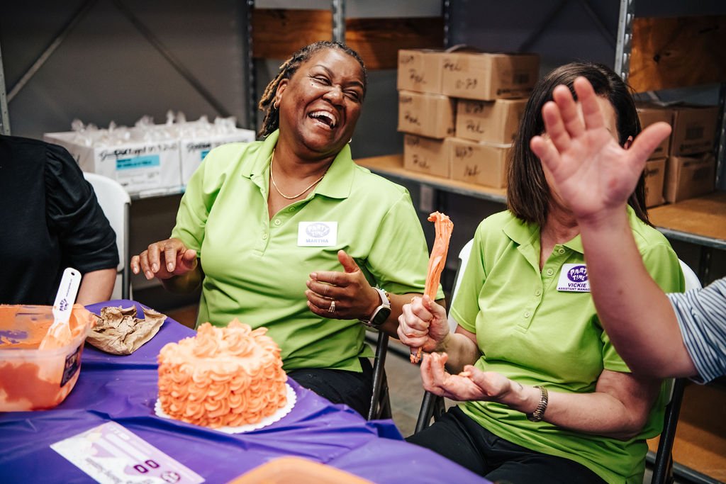 Two women in green shirts laughing and celebrating with a decorated birthday cake and crab legs at a party.