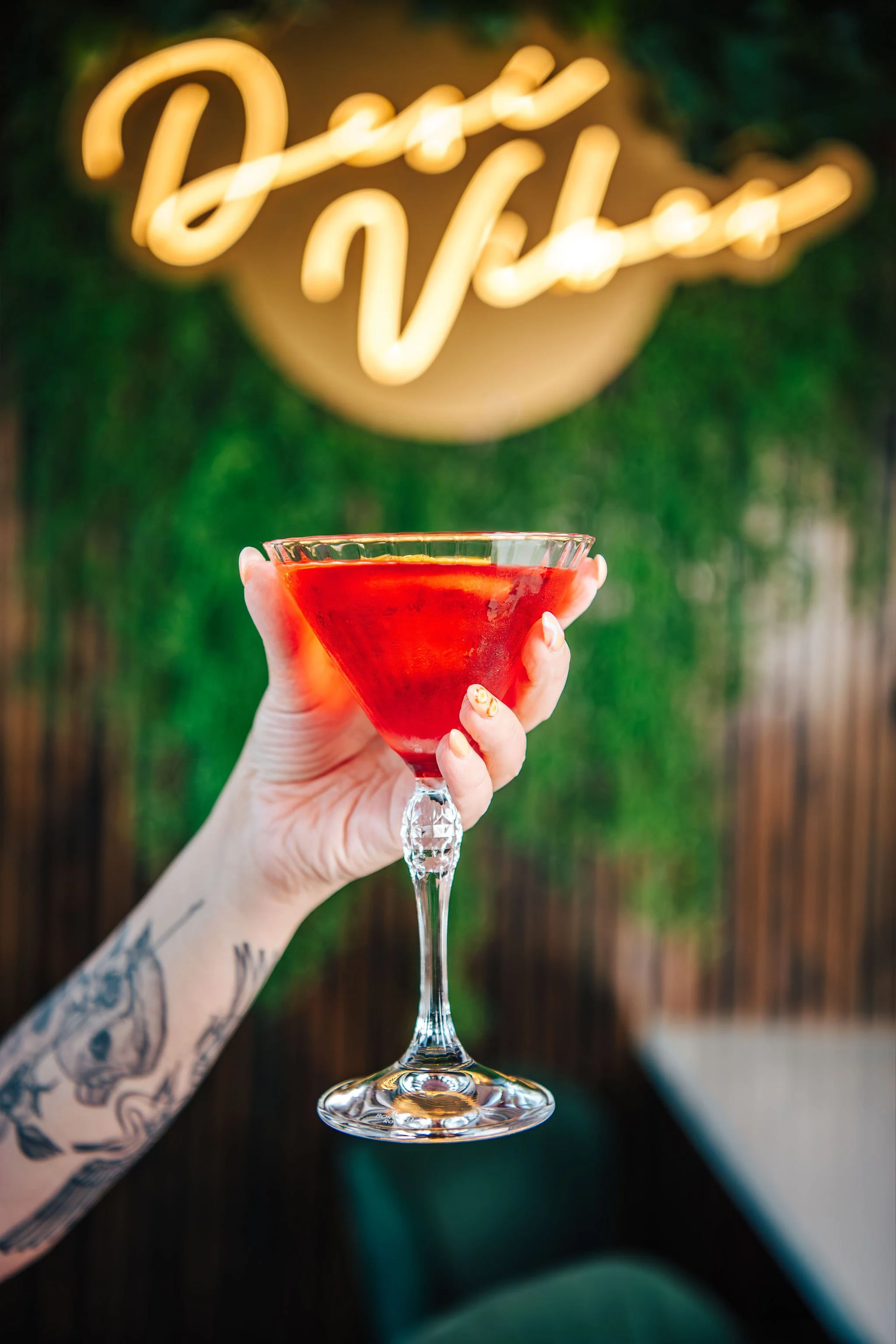 A hand with tattoos holding a glass of red cocktail with a neon sign in the background reading 'Deep Vibes'