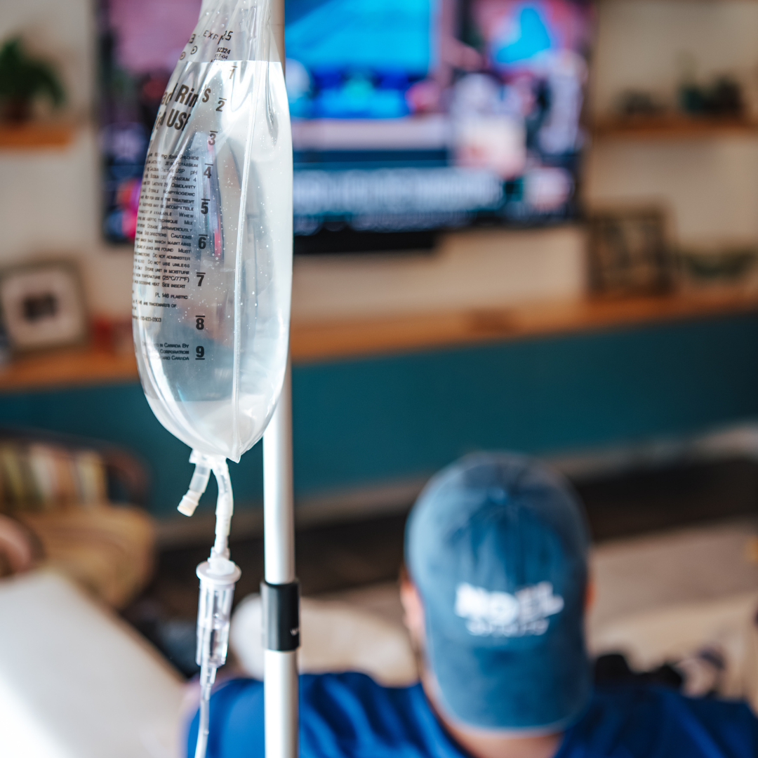 An IV drip with clear liquid hanging in a room, with a person lying on the floor and watching TV in the background.