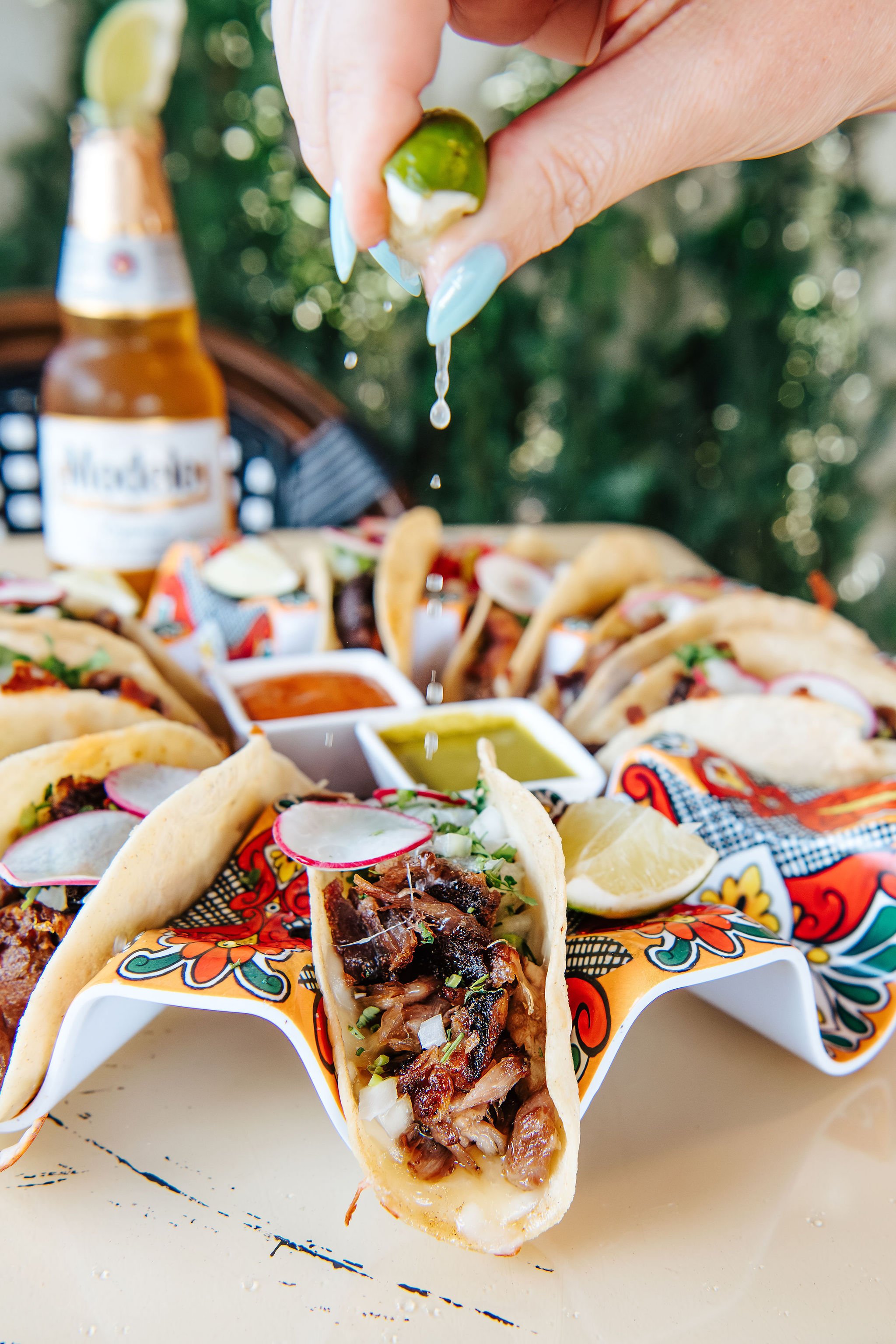 Hand squeezing lime juice over a tray of assorted tacos with dipping sauces on a table.