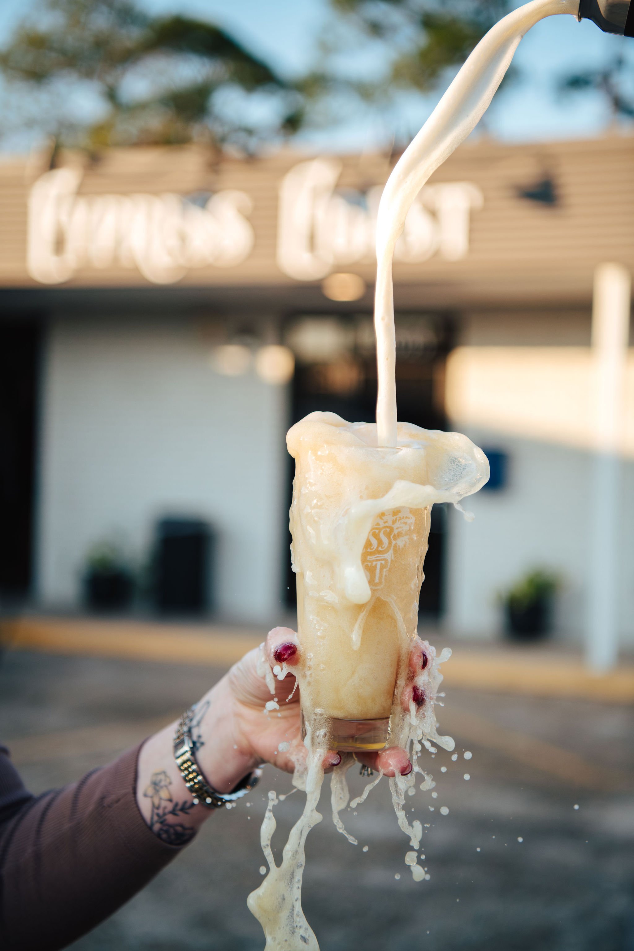 A hand holding a plastic cup with a coffee beverage overflows with cream, causing it to spill and splash. In the background, there's a blurry building with a sign that looks like 'PRES LOCK'.