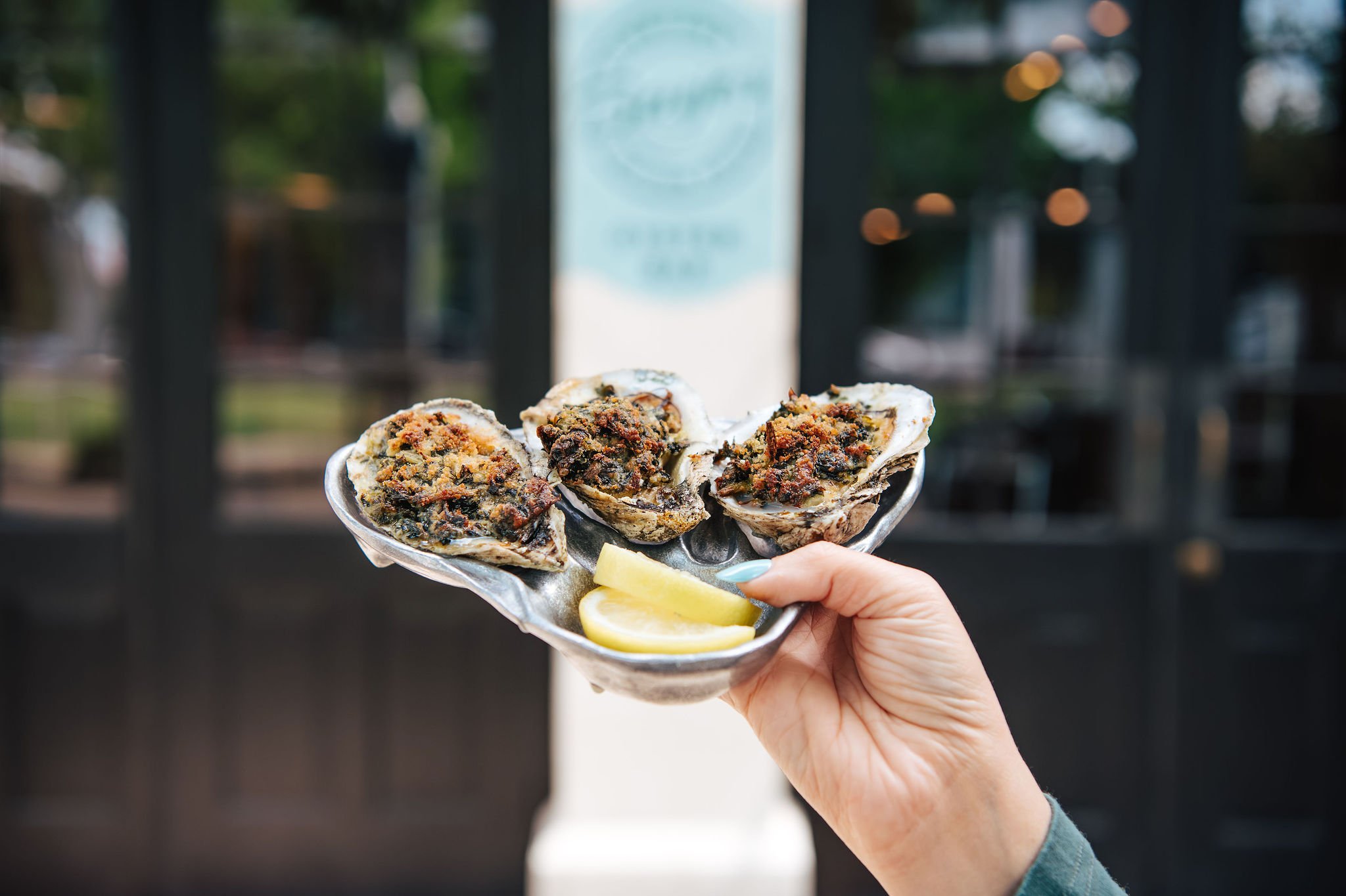 Hand holding a silver dish with three baked oysters topped with seasoning and two lemon wedges in front of a dark outdoor background.
