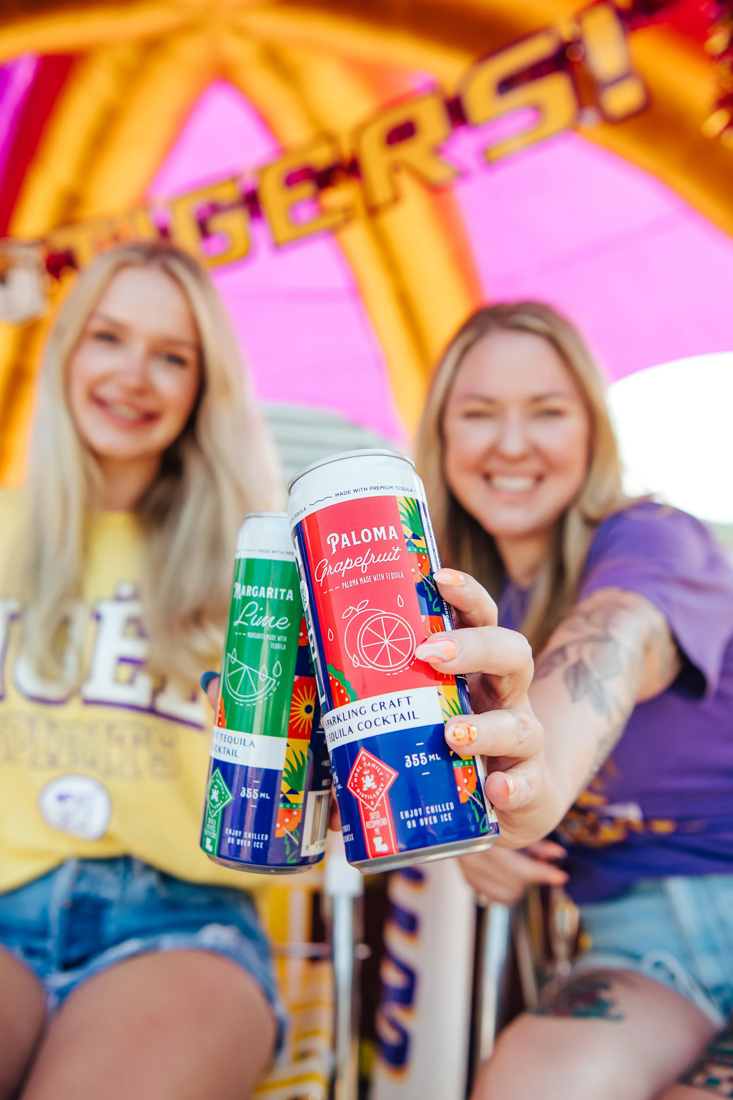 Two women sitting on a carnival ride holding colorful cans of tequila cocktails, with the word 'TERROR' visible on the ride's top. The cans are labeled 'Paloma Grapefruit' and 'Margarita Lime,' with vibrant designs.