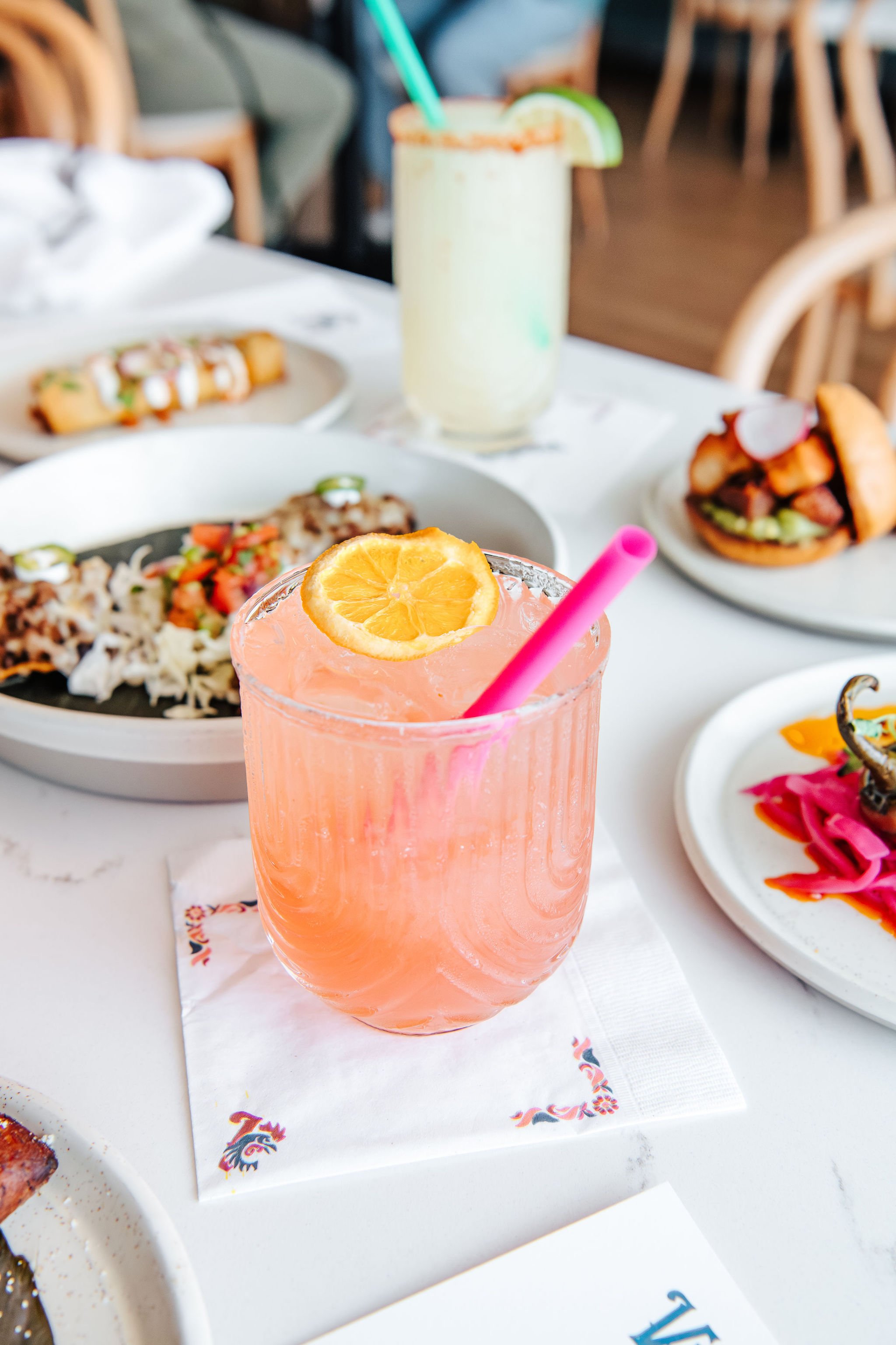 A pink beverage with a lemon slice and pink straw on a white table, surrounded by plates of food including tacos, a burger, and a colorful appetizer.