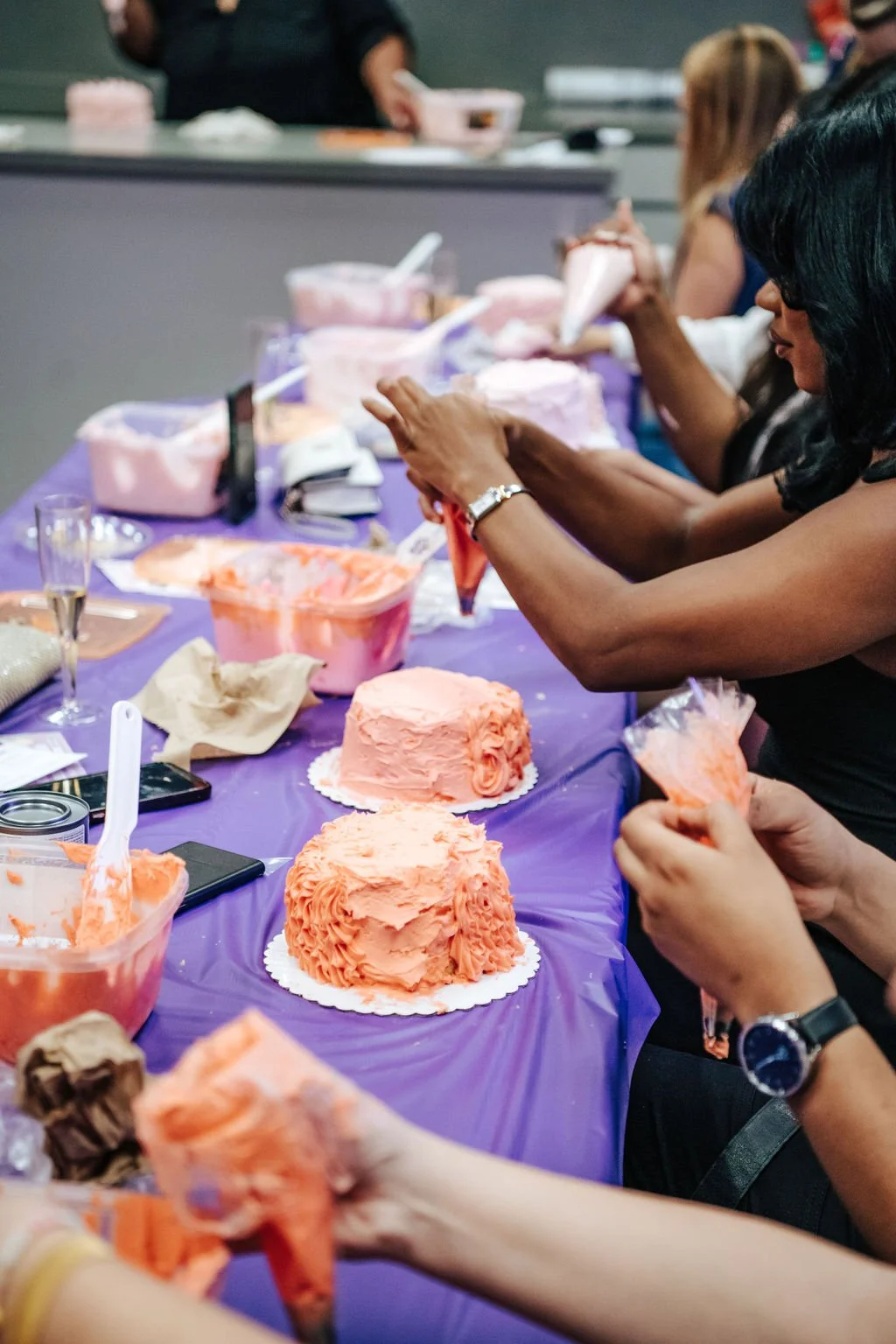 Women decorating pink frosted cakes at a party with a purple tablecloth.