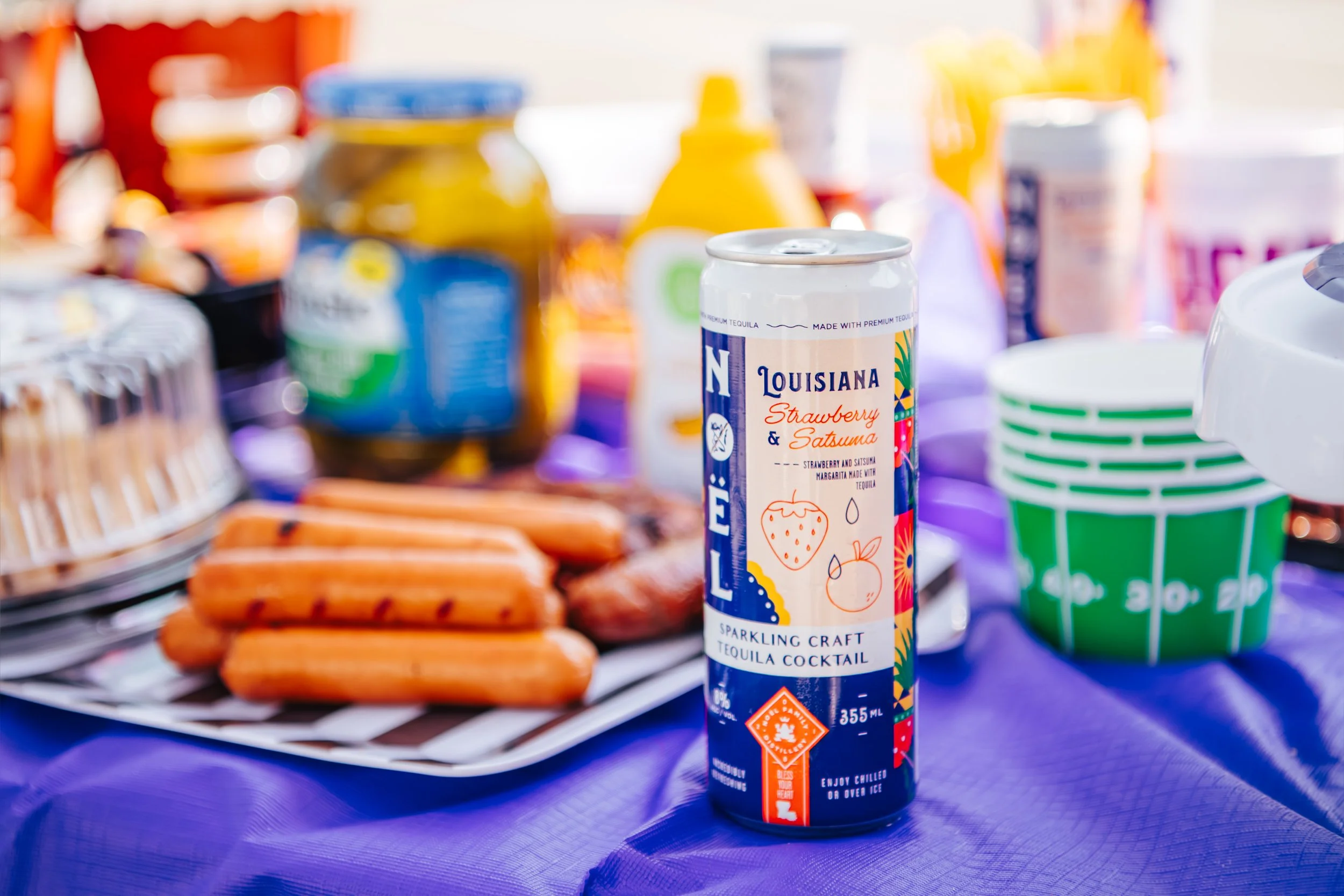 A can of Louisiana Strawberry & Satsuma flavored tequila cocktail on a table with hot dogs, snacks, jars, and colorful cups in the background
