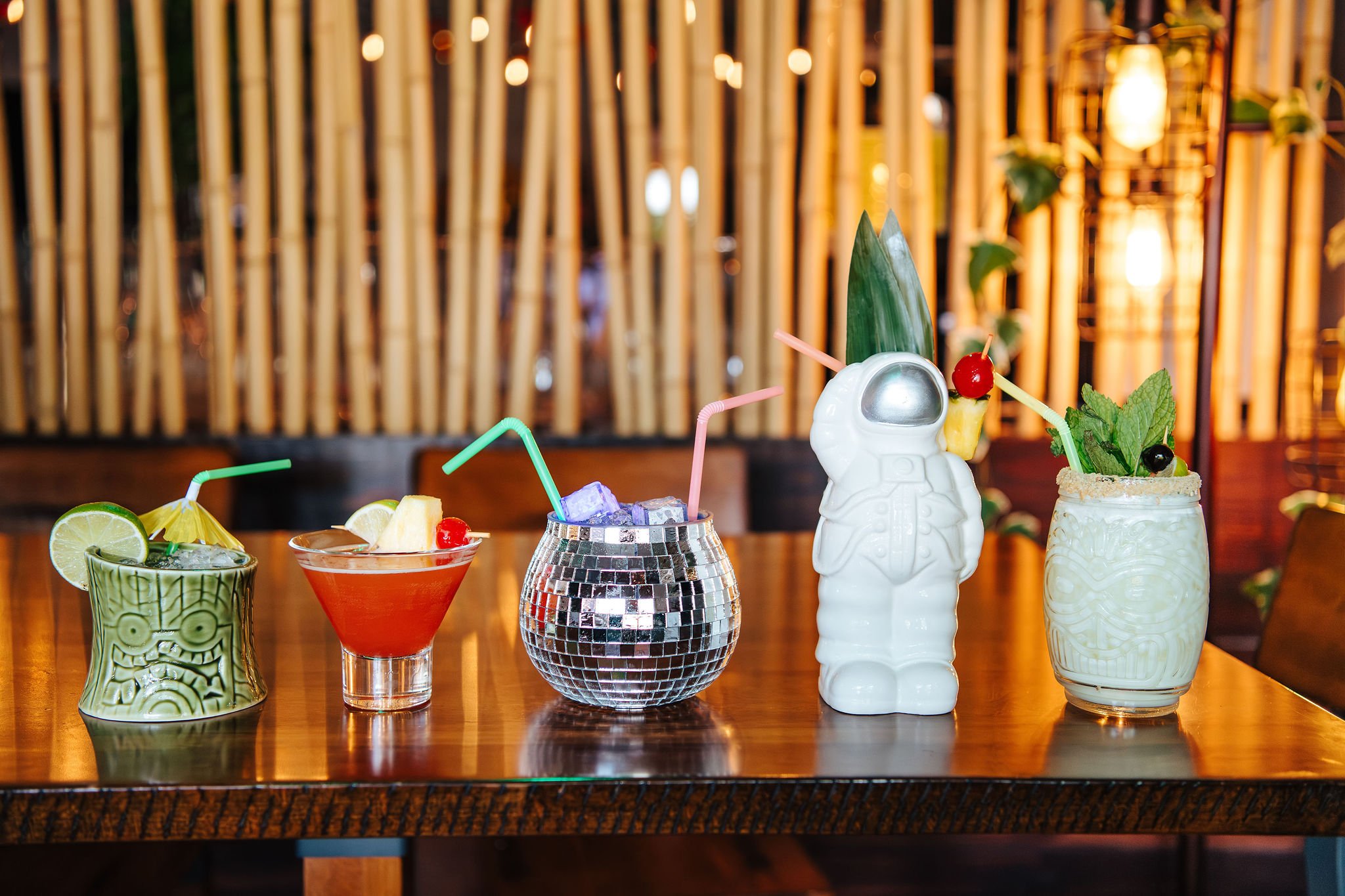 Five colorful tiki-style drinks on a wooden table in a tropical-themed bar with bamboo and warm lighting.