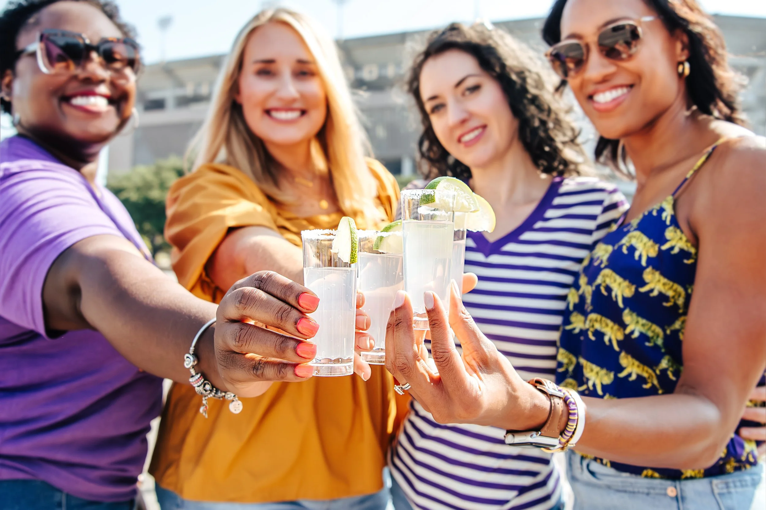 Group of four women toasting with margarita glasses garnished with lime slices outdoors on a sunny day.