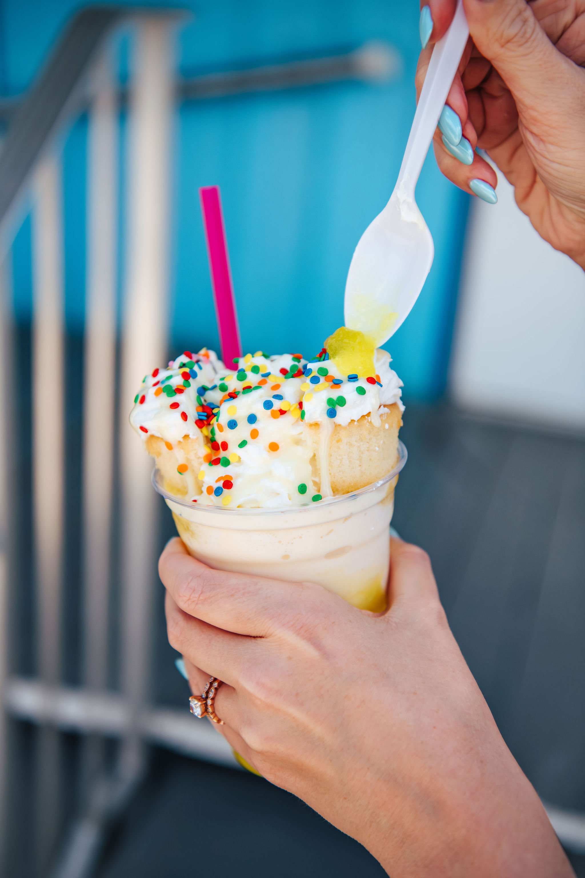 A person with light blue painted nails holding a clear plastic cup with a vanilla milkshake topped with whipped cream and colorful sprinkles. The milkshake has a pink straw, and there is a spoon inserting into the topping, which appears to be a piece