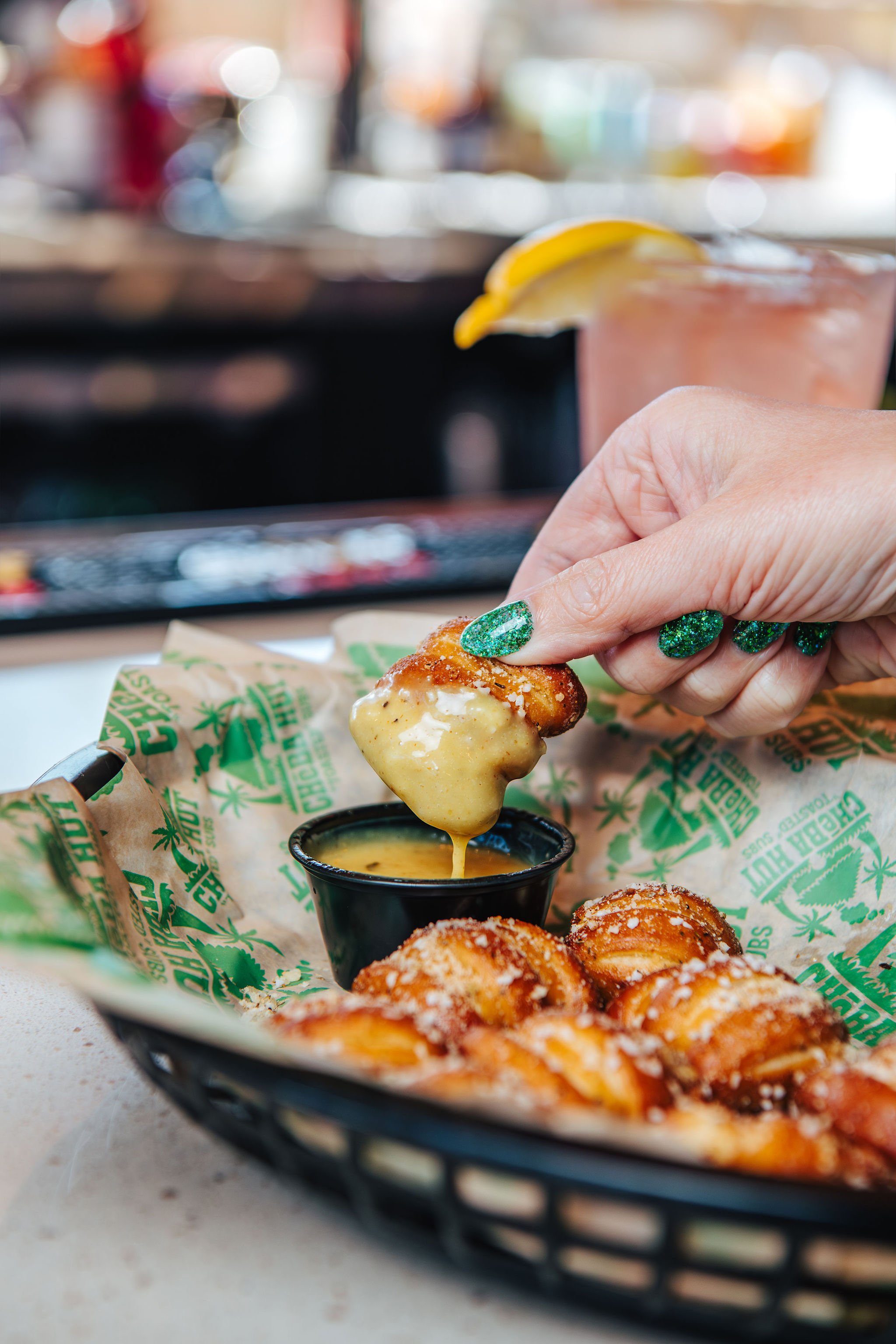 A hand with glittery green nail polish dips a fried cheese curd into a cup of cheese dip, with a basket of fried cheese curds and a pink drink with a lemon wedge in the background.
