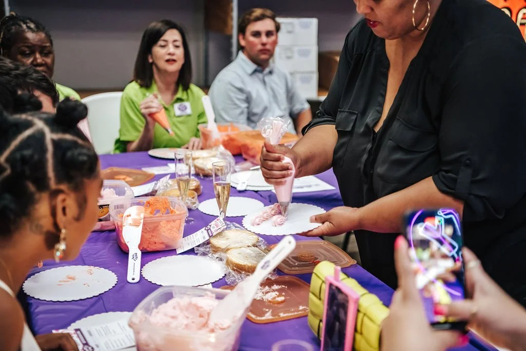 A group of people gathered around a table decorated with a purple tablecloth, watching a woman pipe pink frosting onto a small cake decorated for a celebration. Several glasses of champagne and other desserts are on the table, and one person is taking a photo of the scene.
