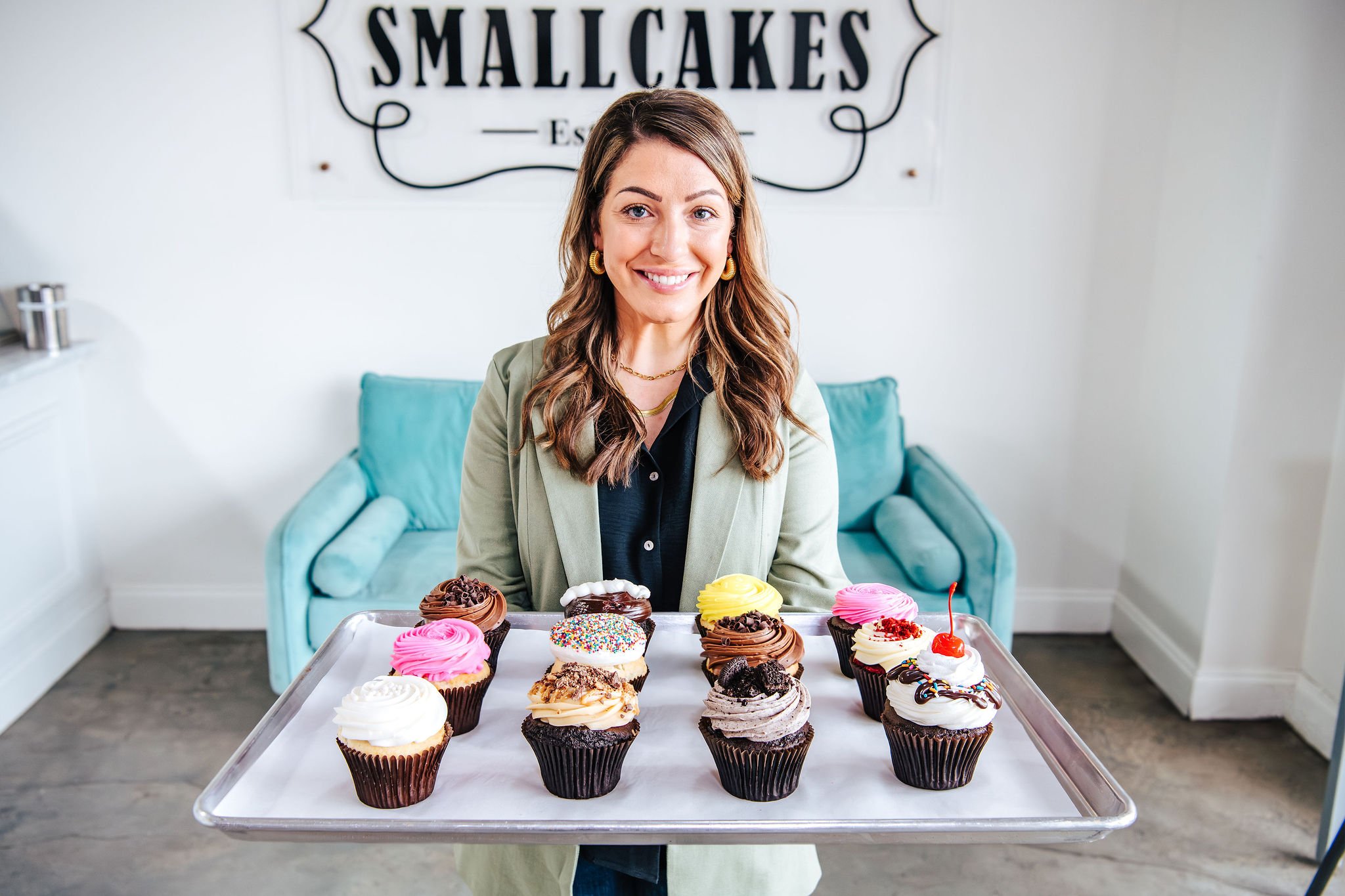A woman holding a tray of assorted cupcakes in a bakery with a sign that reads "Smallcakes" in the background.