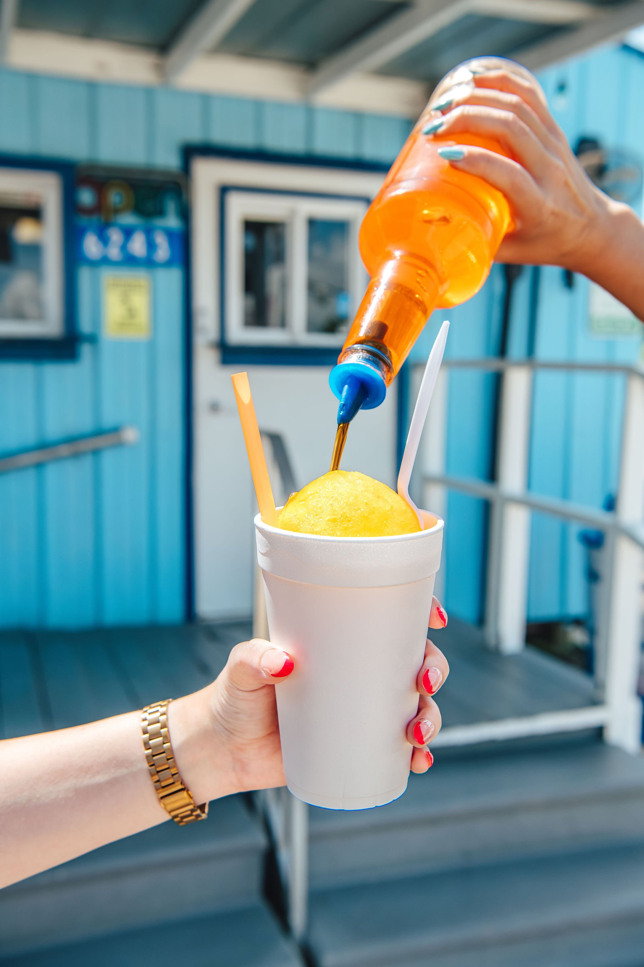 A hand with a gold watch holding a white styrofoam cup with a yellow slush and two straws, while a woman’s hand is pouring orange syrup or juice into the slush from a plastic bottle.