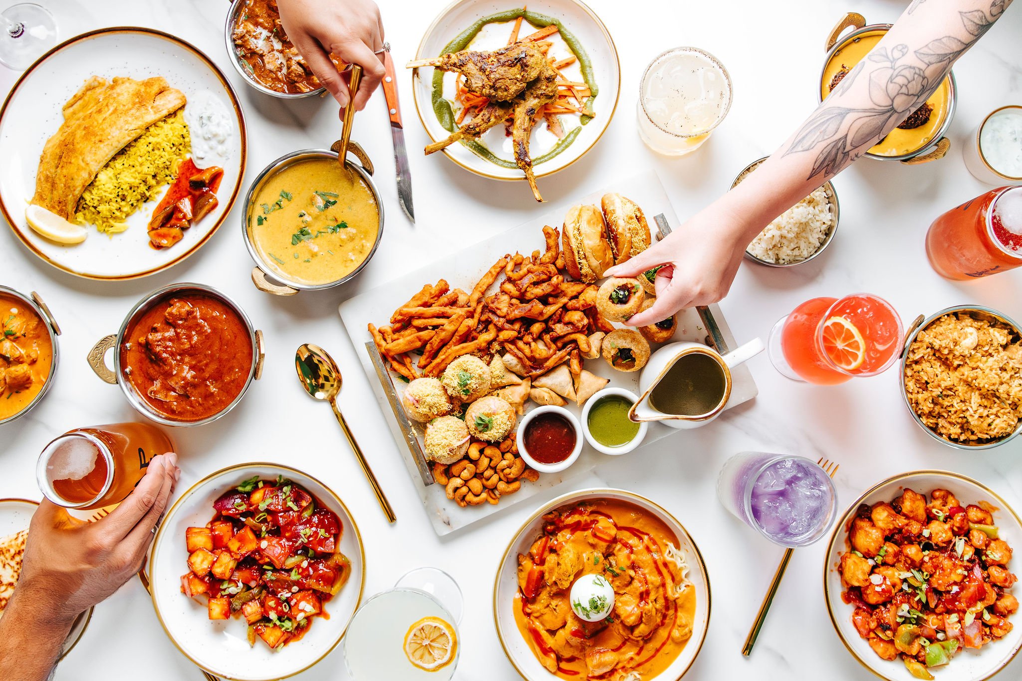A variety of Indian dishes and drinks on a white table, including curry, rice, fried snacks, and beverages.