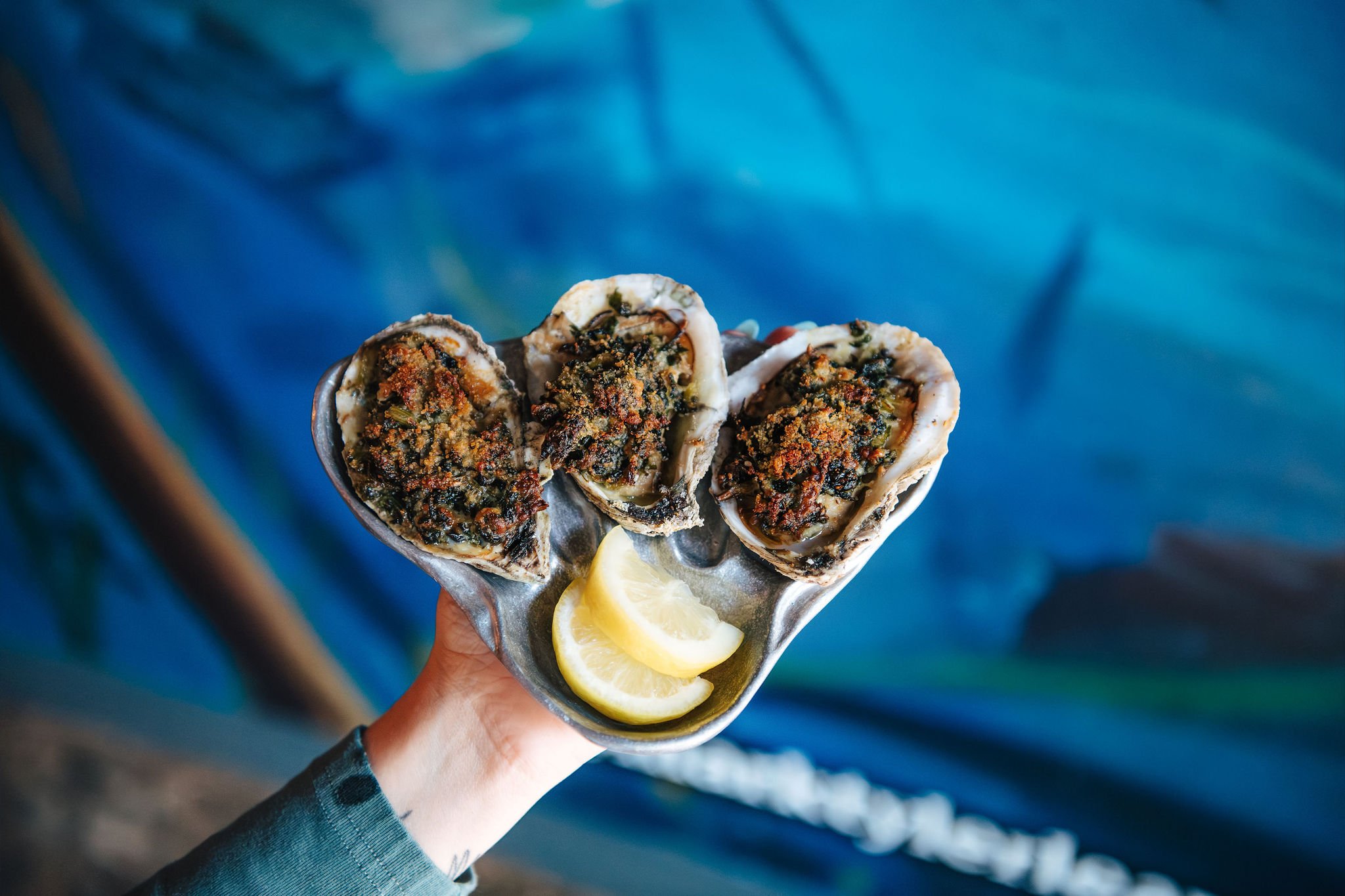 A hand holding a metal plate with three cooked oysters topped with herbs and seasoning, and two lemon wedges on a blue background.