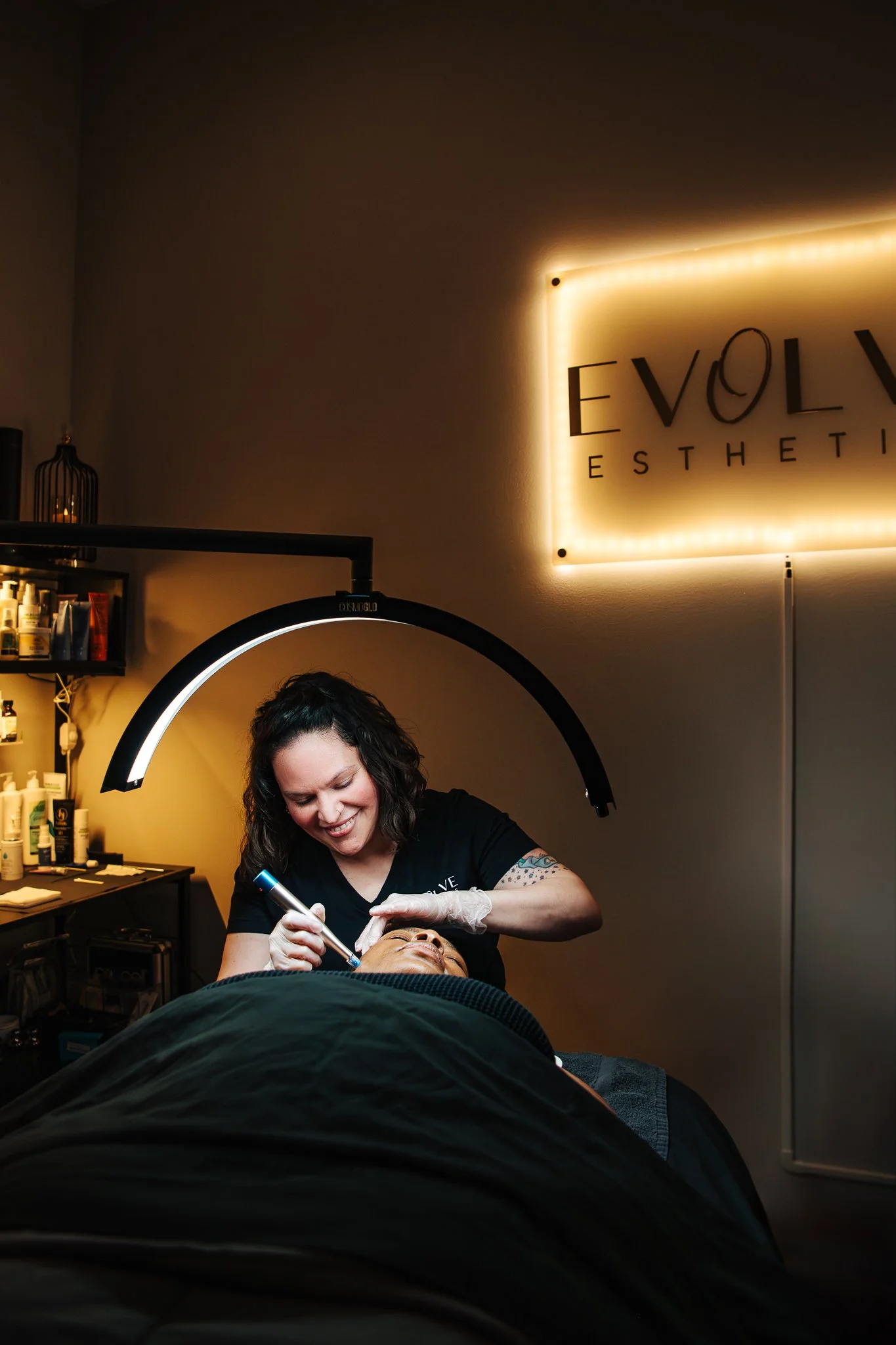 A woman performing a cosmetic procedure on a patient in a modern esthetic clinic with a sign that reads 'Evolve Esthetics.'
