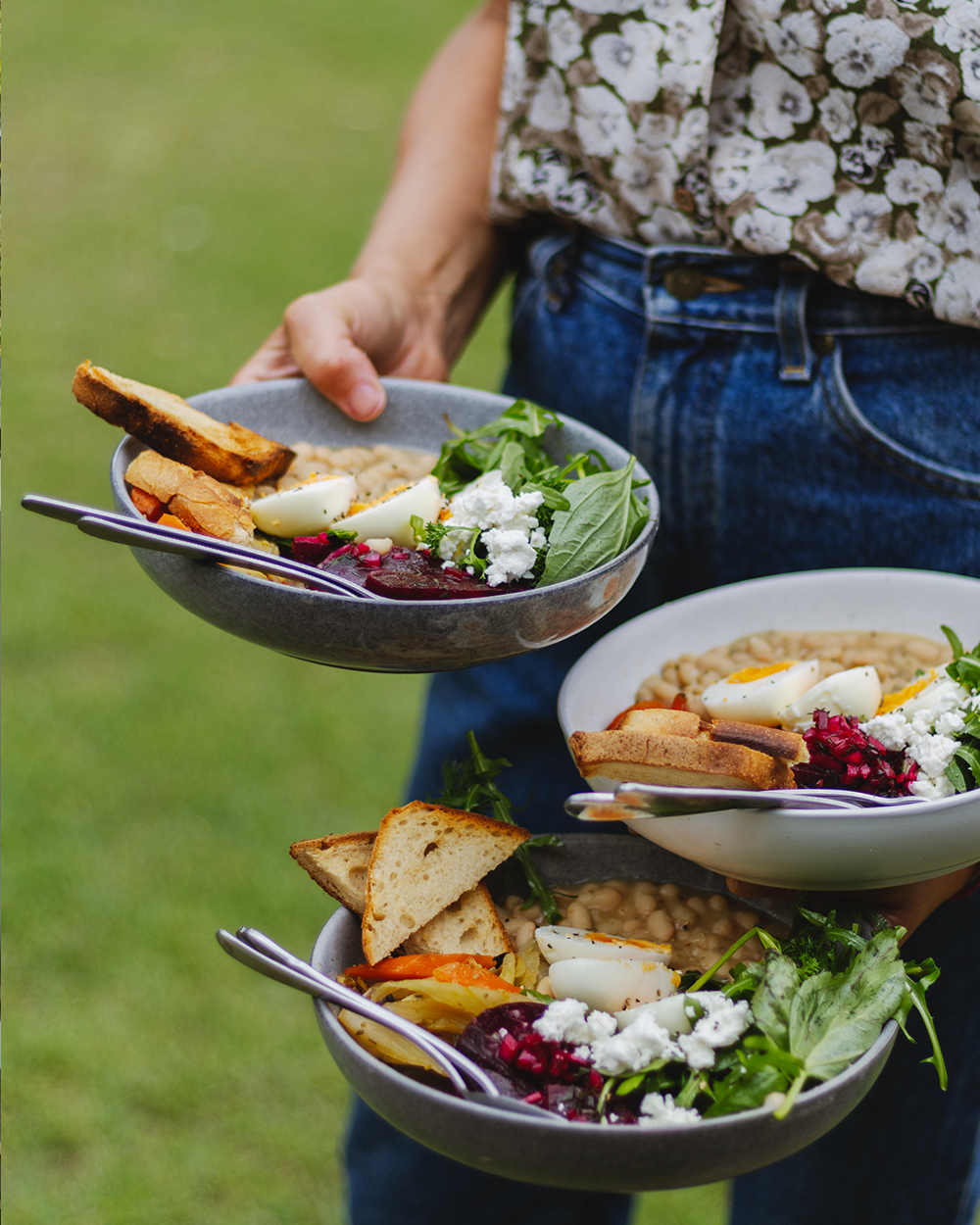 Person holding three bowls of salad with boiled eggs, bread, greens, and beets outdoors.