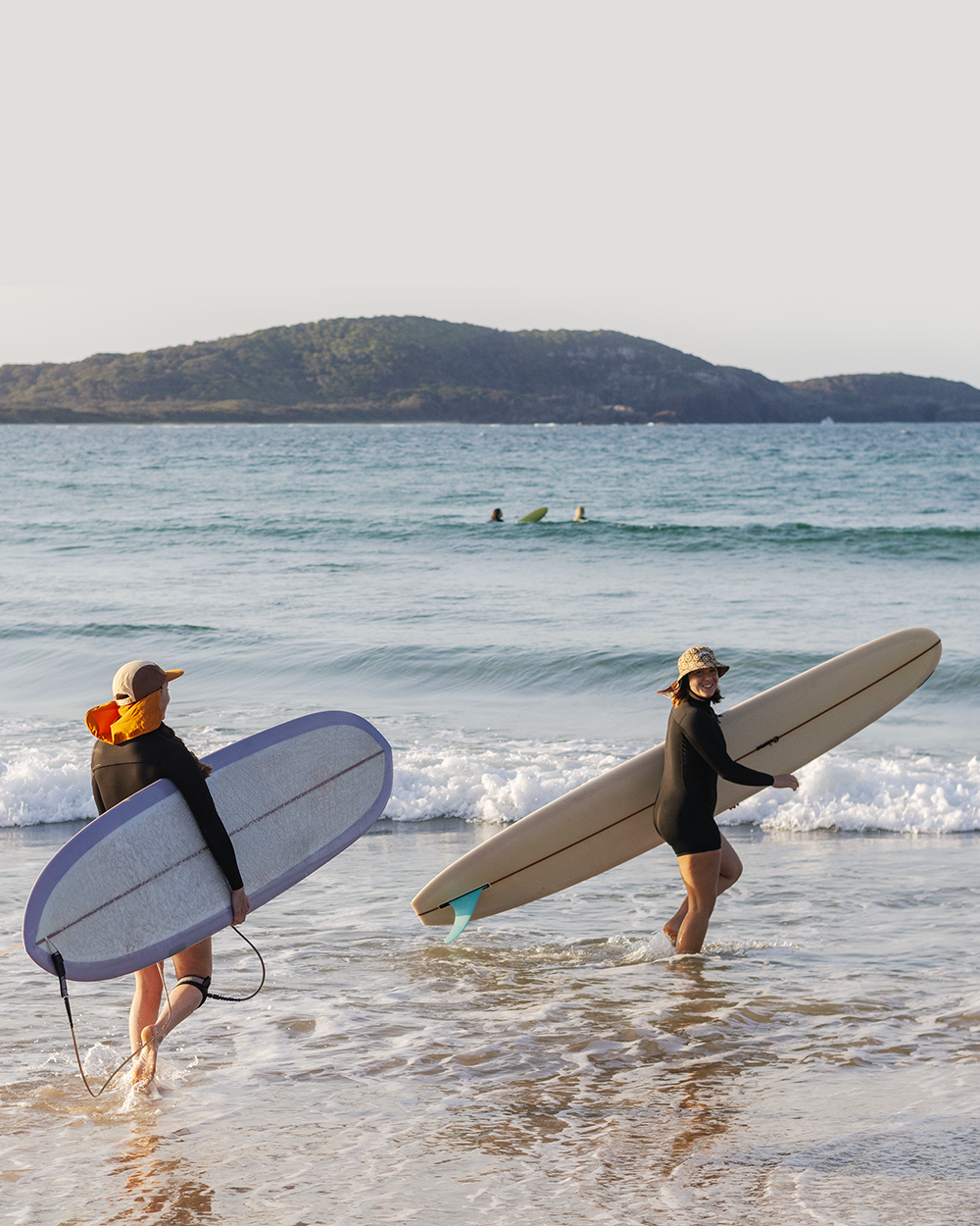 Two women carrying surfboards walk out of the ocean with a hill in the background.