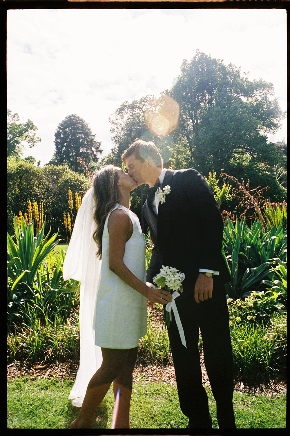 A bride and groom kiss outdoors in a garden, with lush greenery and trees in the background, as sunlight shines through the trees.