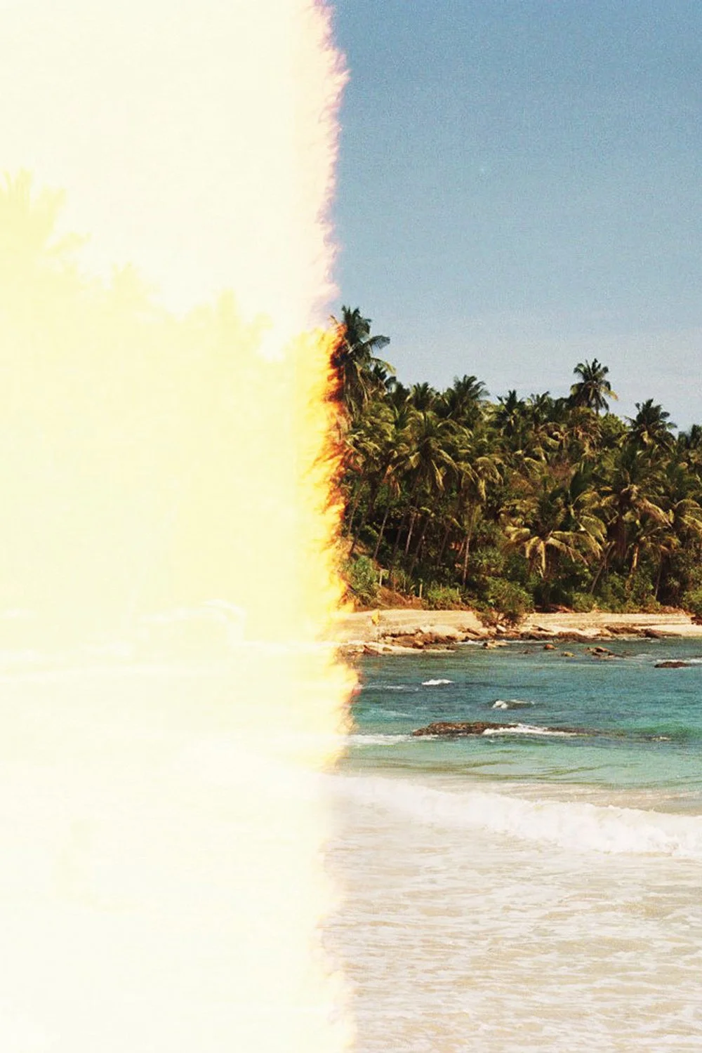 Photo of a tropical beach with palm trees, ocean waves, and a clear sky, partially obscured by light leak or overexposure on the left side.