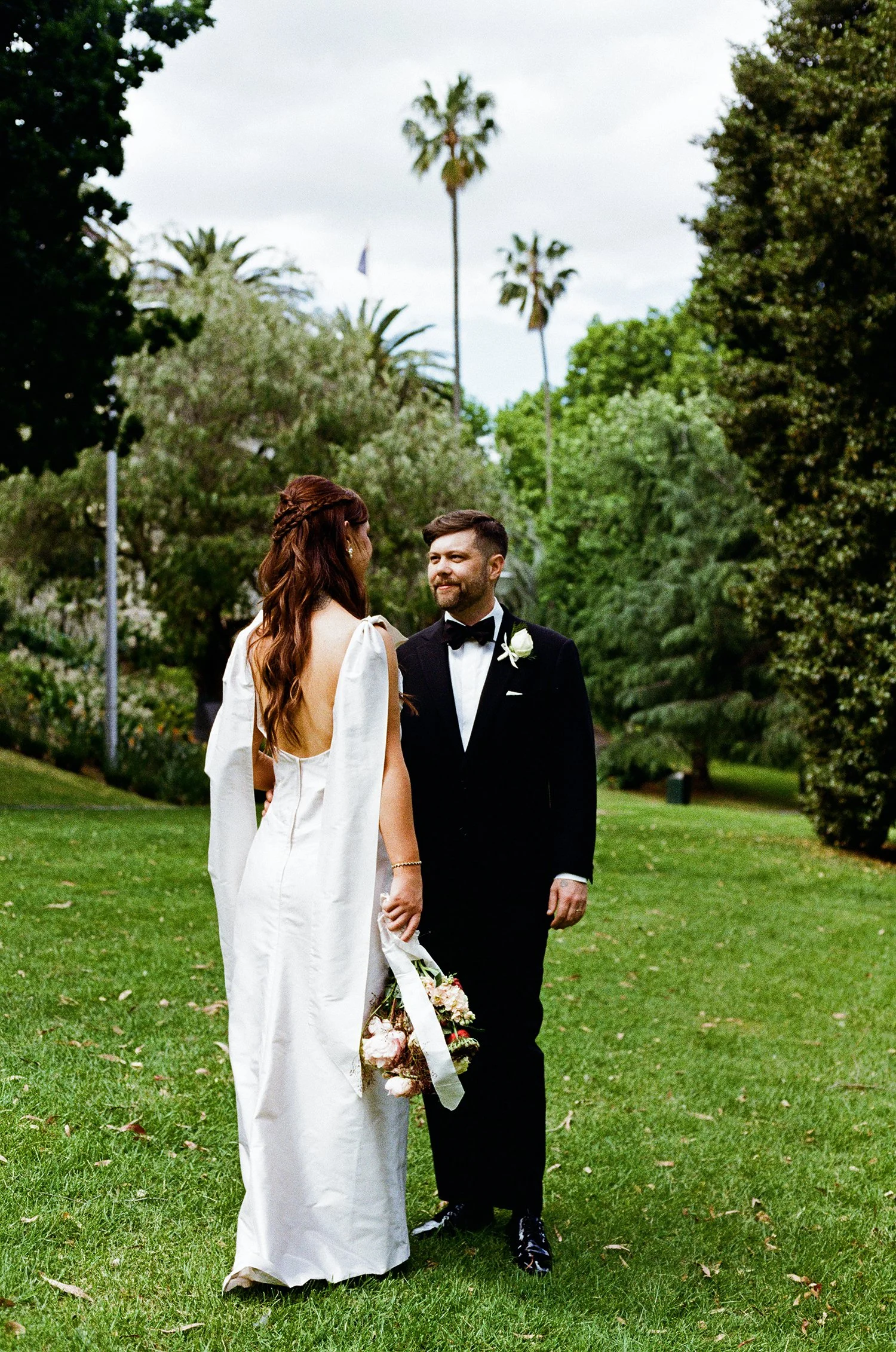 A bride and groom standing on green grass in a park with trees and palm trees in the background. The bride is holding a bouquet and wearing a white wedding dress with open back and puffed sleeves. The groom is wearing a black tuxedo with a bow tie an
