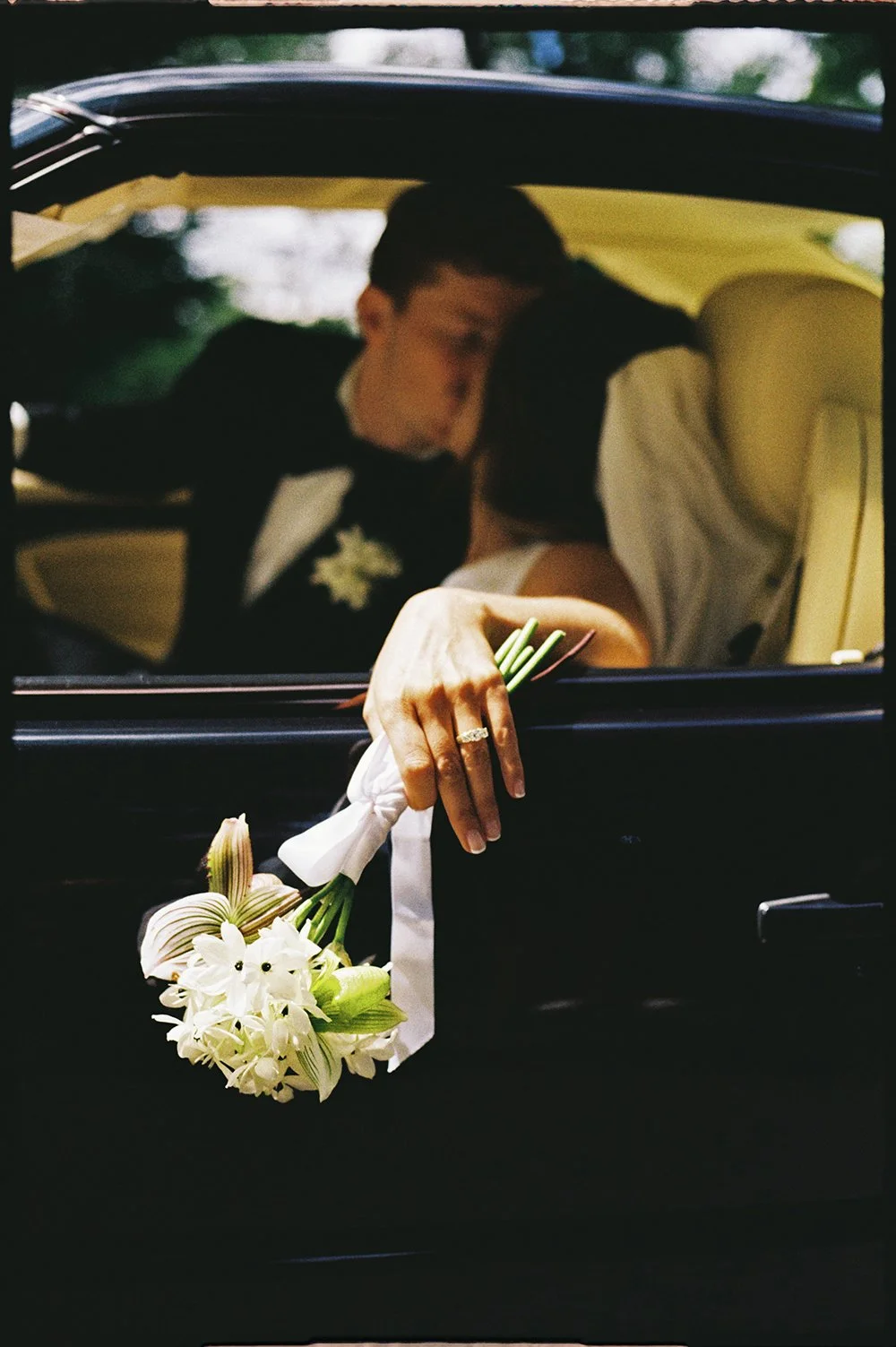 A bride and groom sharing a kiss inside a car, with the bride holding a bouquet of white flowers hanging outside the window.