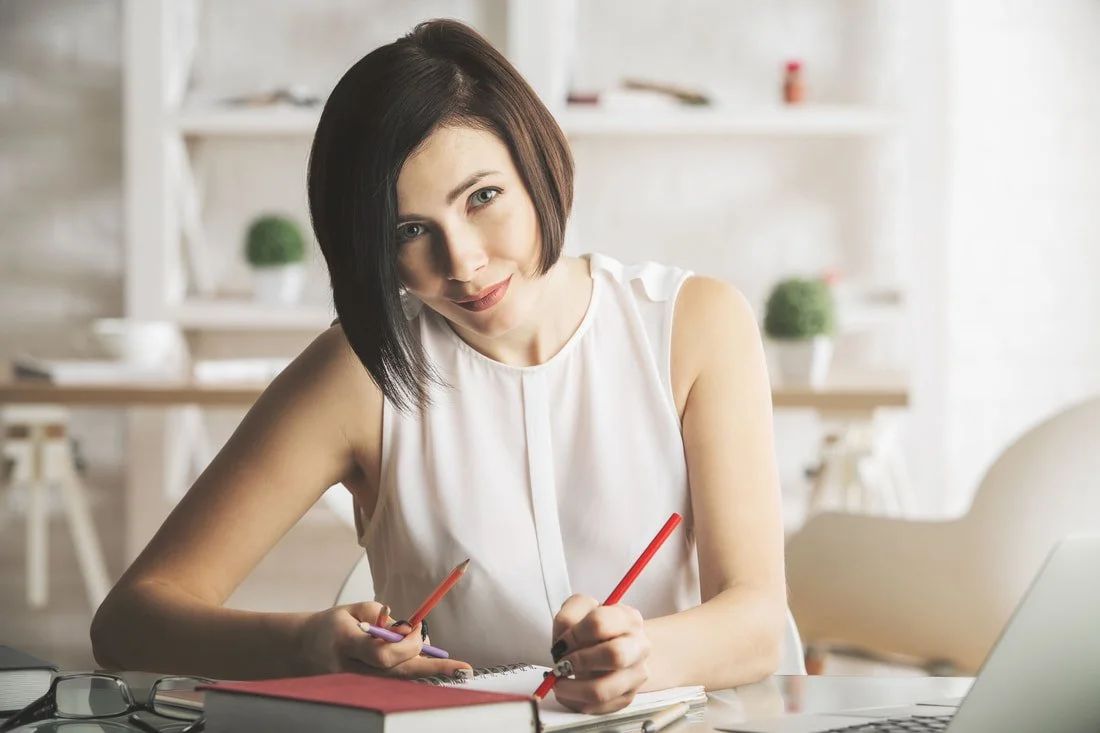 A woman with short dark hair sitting at a desk with notebooks, pens, and a laptop in a well-lit room.