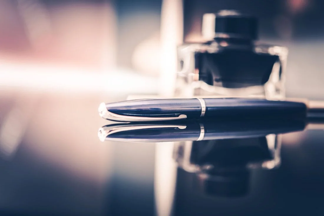 Close-up of a black fountain pen lying on a reflective surface with a blurred perfume bottle in the background.