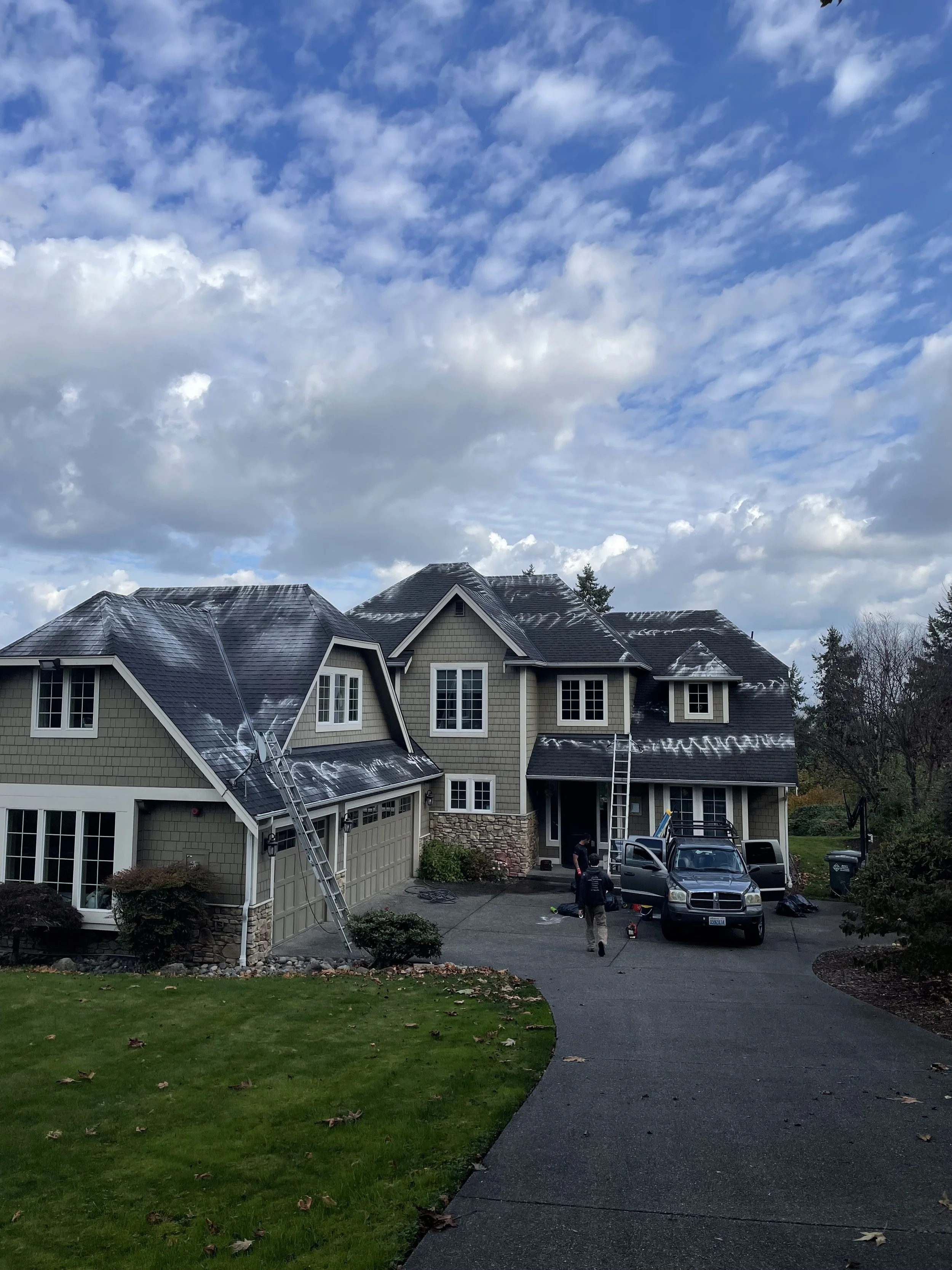 A house with a dark gray shingle roof under a partly cloudy sky. Two ladders and workers are on the roof, appearing to perform maintenance. The driveway has trucks and tools, and there's a lush green lawn in the front yard.