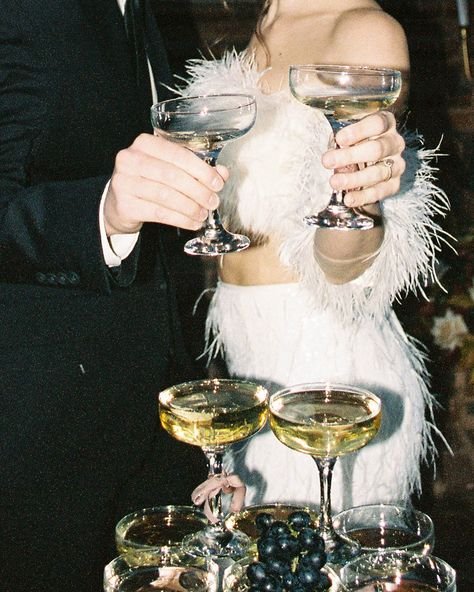 A woman in a white feathered dress holding two champagne glasses, standing next to a man in a dark suit holding two champagne glasses, with a table of wine glasses and grapes in the foreground.
