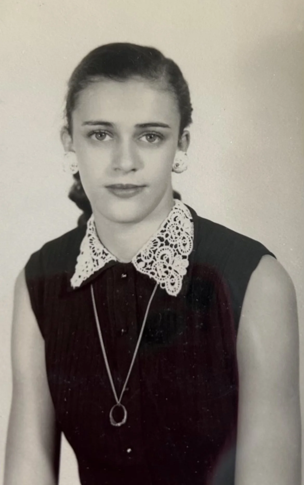 Black and white portrait of a young woman wearing a sleeveless dark dress with a lace collar, earrings, and a necklace, looking at the camera with a neutral expression.