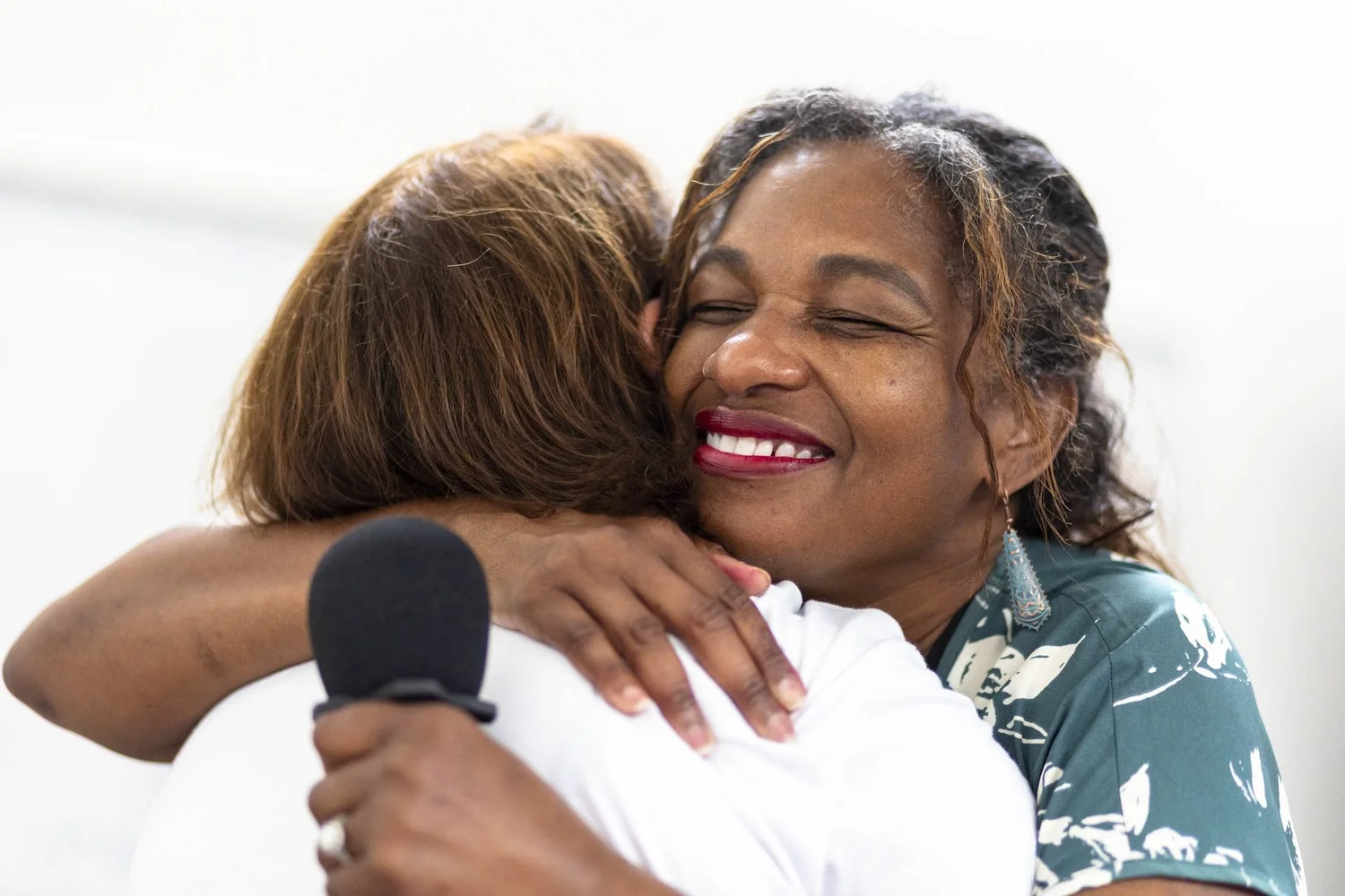 Two women share a warm hug at a training, one smiling with her eyes closed and holding a microphone