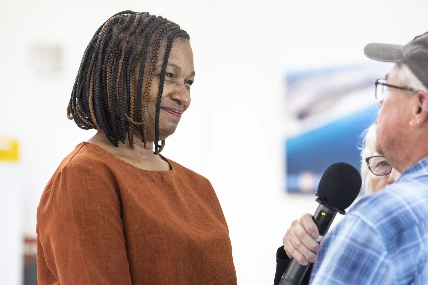 A woman with braided hair smiles as she listens, while a man in the foreground holds a microphone